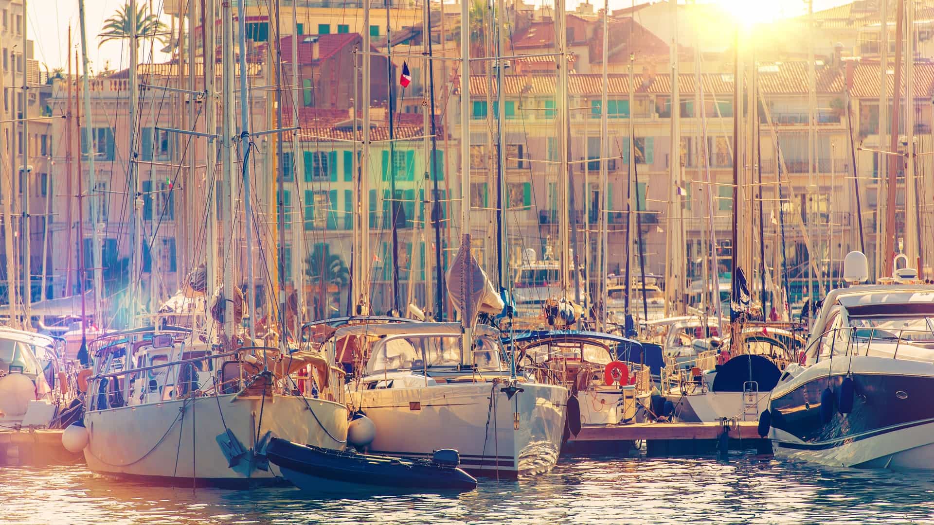 A scenic view of the Cannes marina on the French Riviera, featuring numerous yachts and boats docked in the harbor. In the background, the buildings of the city of Cannes are visible under a clear sky.