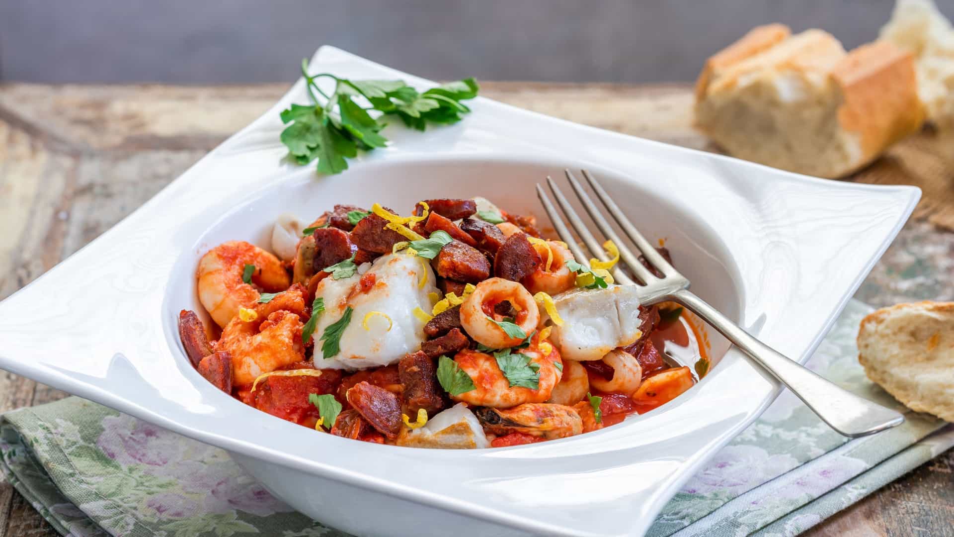A close up, top down view of a bowl of bouillabaisse, a traditional French fish stew. The rich, red orange broth is filled with chunks of fish and seafood. It is garnished with fresh herbs and slices of crispy chorizo, and served with a piece of toasted bread on the side.