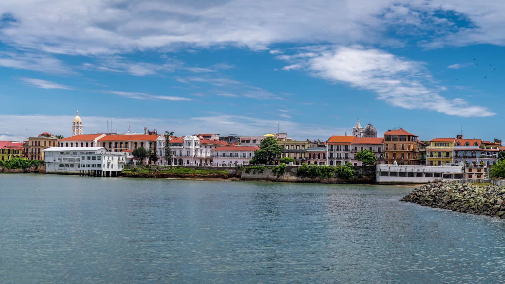 A panoramic aerial view of Casco Viejo, the historic district of Panama City, Panama. The image shows a dense collection of colorful colonial era buildings with red tile roofs, narrow streets, and historic plazas. In the background, the modern skyline of downtown Panama City rises, with towering skyscrapers reflecting the sunlight, creating a striking contrast between the old and the new.