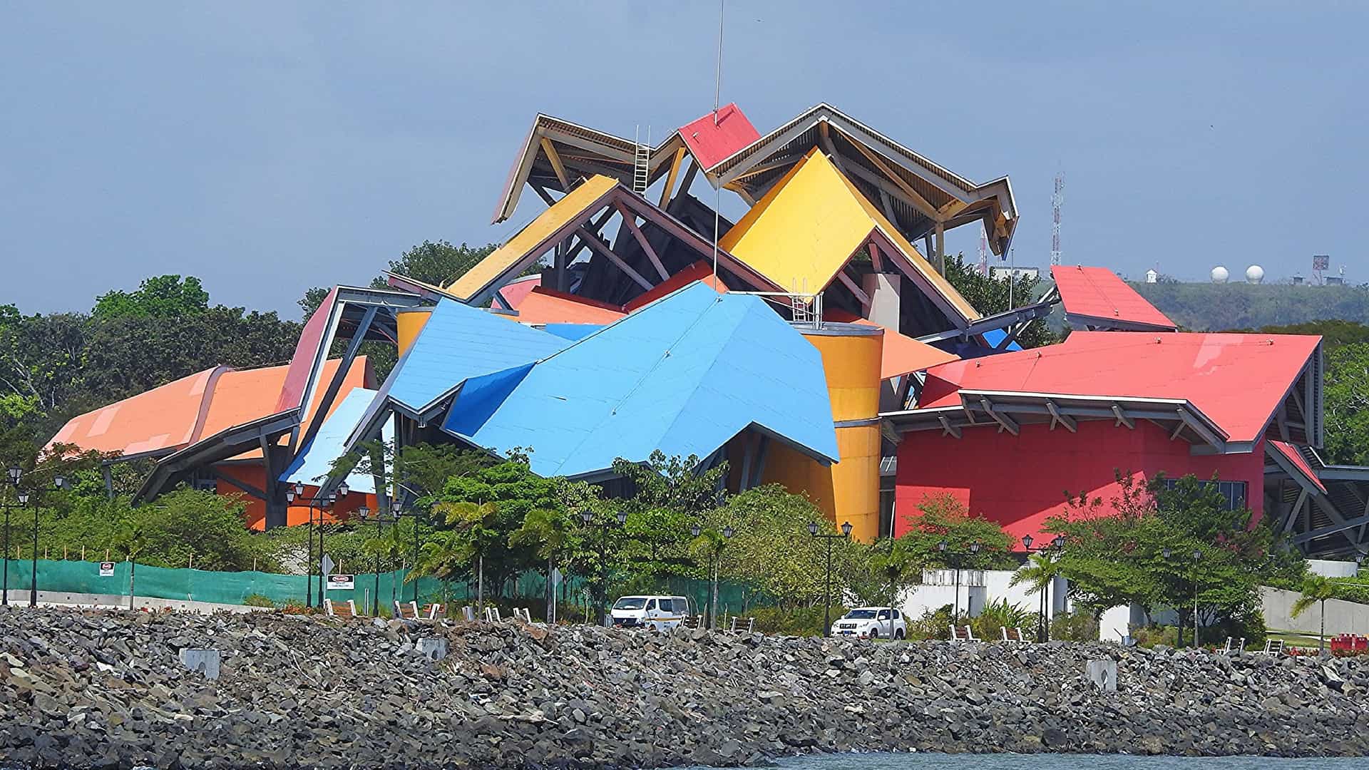A close-up view of the Biomuseo in Panama City, designed by architect Frank Gehry, on the Amador Causeway. The museum is immediately recognizable by its vibrantly colored, deconstructed roof canopy, which features bold, zigzagging panels in shades of red, blue, yellow, and green. The foreground shows the structure surrounded by lush green trees, with the modern, low-lying museum building visible beneath the striking roof. The design highlights the museum's focus on Panama's incredible biodiversity and its role as a land bridge between two continents.