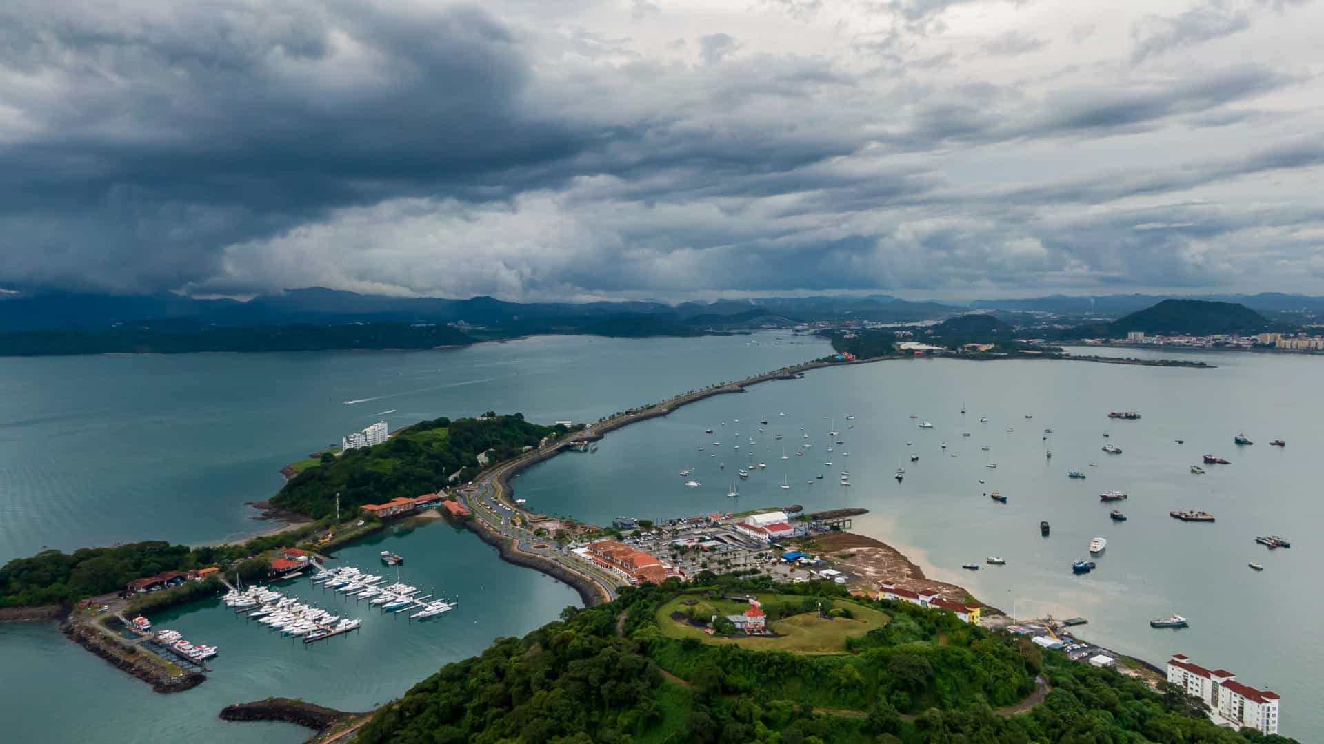 A high angle aerial view of the Amador Causeway in Panama City, Panama. The causeway is a long, curving strip of land connecting the mainland to several small islands in the bay. The image shows the road lined with palm trees, and the blue water of the bay surrounding it. In the distance, the modern skyline of Panama City is visible, creating a contrast between the man made land bridge and the bustling metropolis.
