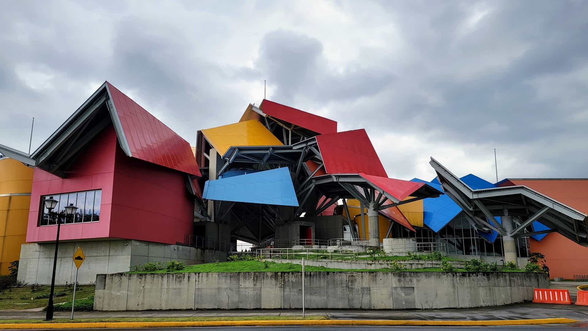 A wide-angle exterior view of the Frank Gehry Bridge of Life Museum (Biomuseo) in Panama City. The museum is instantly recognizable by its complex, multi-layered roof canopy, featuring bold, geometric planes painted in vivid colors like red, blue, yellow, and green. The chaotic, deconstructed roofline is set above the lower, white museum structure, highlighting the unique architectural design against the surrounding lush environment.