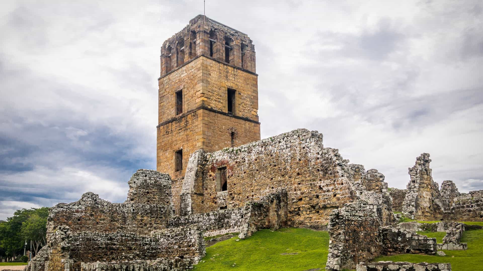 The stone ruins of the old Panama Viejo cathedral and its bell tower stand prominently in a green field under a clear blue sky, with modern high rise buildings of the new Panama City visible in the distance, creating a striking contrast between past and present.