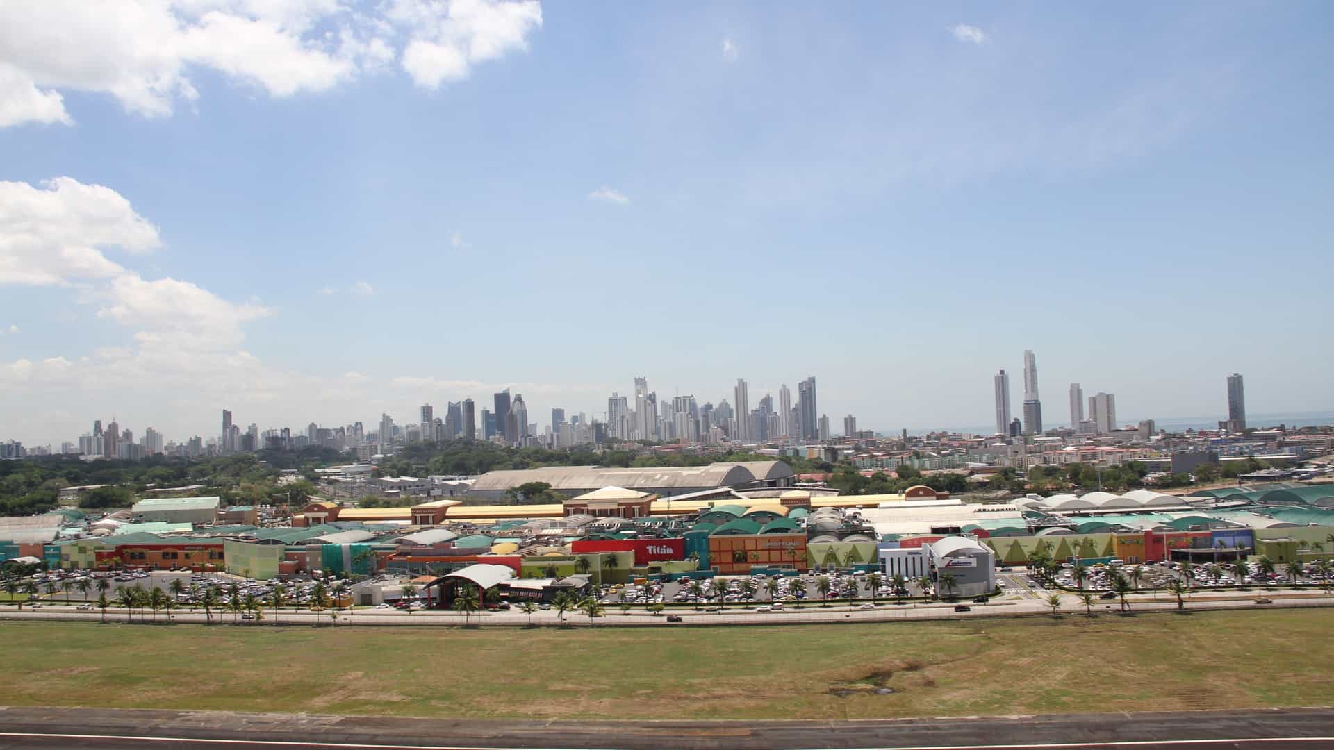 A wide-angle, panoramic view of the Panama City skyline on a bright, sunny day. In the background, the dense cluster of modern, high-rise skyscrapers defines the city's downtown core. The middle ground is dominated by the sprawling, low-rise commercial structure of the Albrook Mall, characterized by its expansive horizontal profile and multi-colored roofing, situated between a grassy area in the foreground and the distant vertical cityscape.