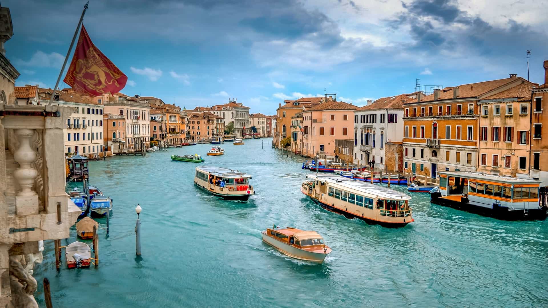 Water taxis and vaporetto boats move along the busy Grand Canal in Venice, with historic buildings and archways lining the banks under a partly cloudy sky.