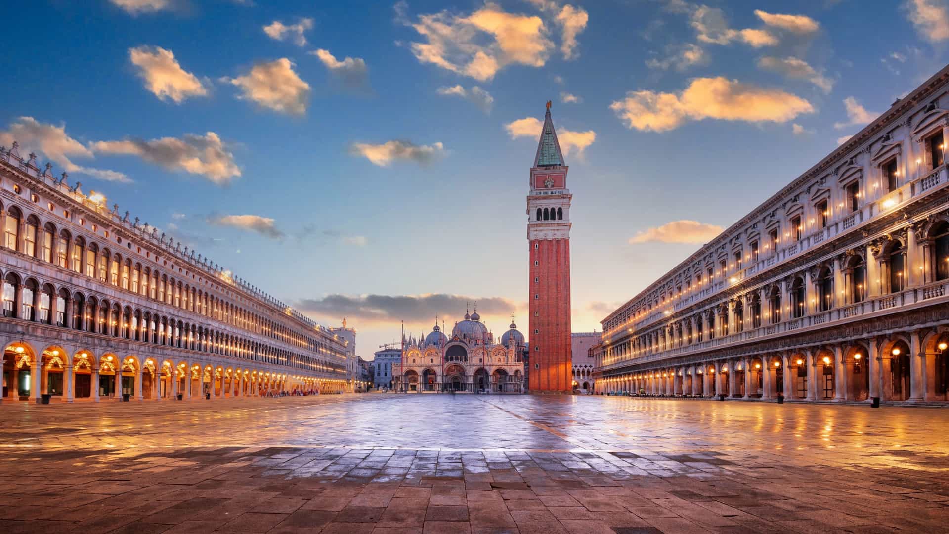 A close up view of the intricate facade of St. Mark's Basilica in Venice, showcasing its multiple domes and ornate arches adorned with mosaics. People are visible in the foreground of the square, and the sky is a vibrant blue.