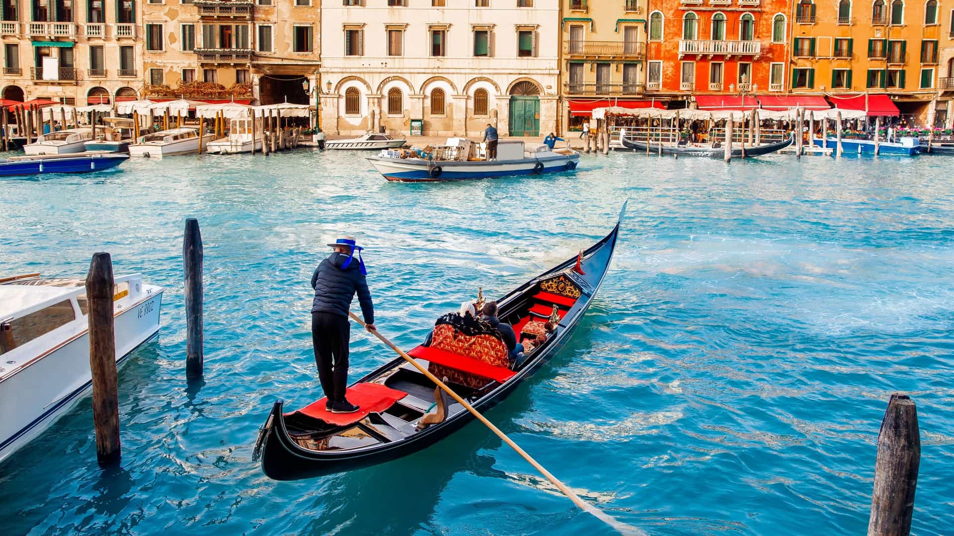 A gondolier, dressed in a traditional striped shirt, stands at the stern of a gondola, navigating tourists through a Venetian canal. The scene features historic buildings lining the waterway.