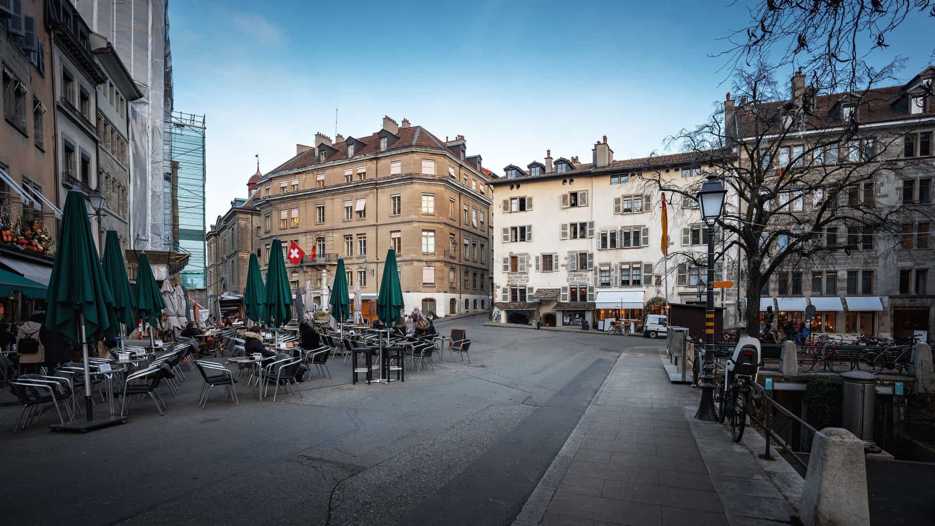 A charming, wide-angle view of the Place du Bourg-de-Four, Geneva's oldest square, in the Old Town. The cobbled, pedestrian square is surrounded by multi-story, 16th to 18th-century European buildings with colorful facades, ground-floor cafes featuring outdoor seating, and small trees, all centered around a prominent, classic stone fountain.