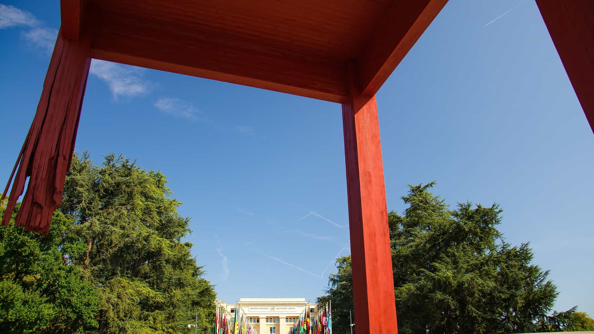 A towering, 12-meter high wooden sculpture known as the "Broken Chair" stands prominently in the Place des Nations in Geneva, Switzerland. The massive chair is missing one of its front legs, symbolizing the opposition to landmines and cluster bombs. In the background, the modern Palais des Nations (United Nations Office) building is visible, with a row of international flags lining the entrance.