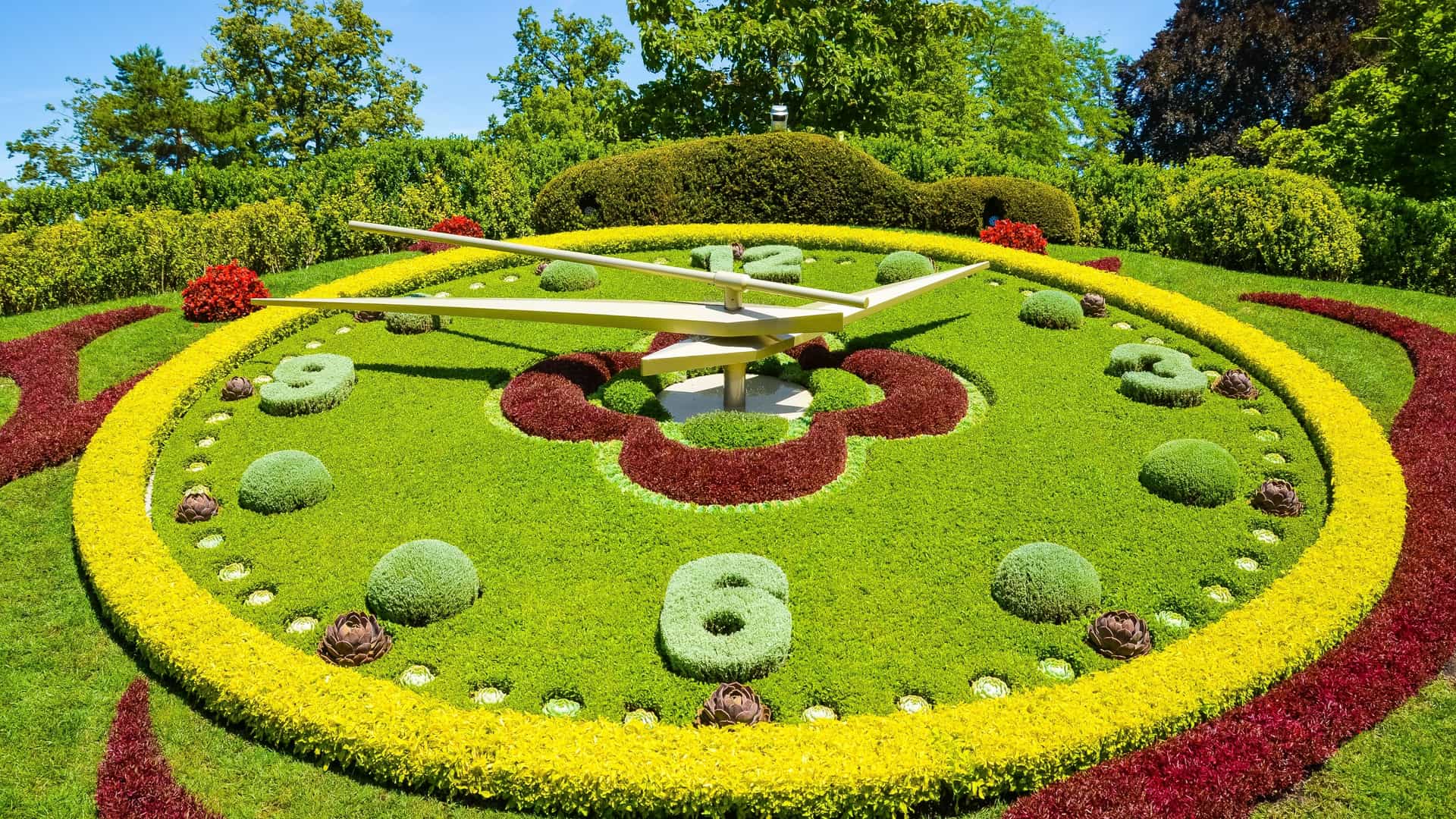 An aerial view of the L'Horloge Fleurie (Flower Clock) in Geneva's Jardin Anglais. The large outdoor clock face, set into a green lawn, is elaborately composed of thousands of brightly colored, meticulously arranged flowering plants and shrubs, featuring golden hands and white markers, symbolizing the city's world-renowned watchmaking tradition.