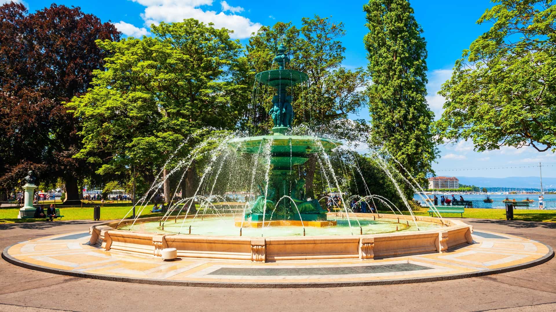 A detailed, eye-level photo of the bronze Fontaine des Quatre Saisons (Fountain of the Four Seasons) located in the Jardin Anglais in Geneva. The ornate, tiered fountain features classical figures and is actively spraying water from its top basin, set against a backdrop of green trees and paved pathways in the park.