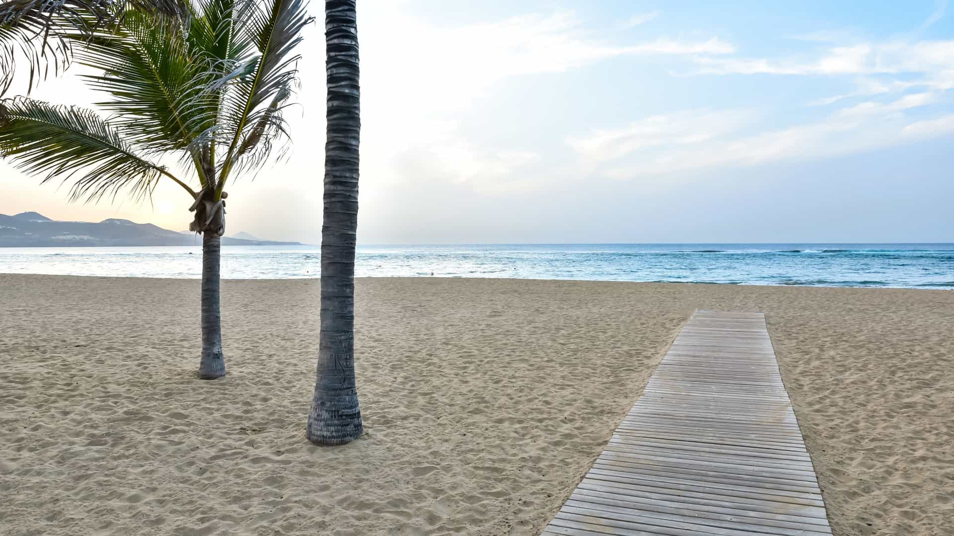 A scenic view of Playa de las Canteras in Las Palmas, Gran Canaria, Spain. The image shows a wide, sandy beach with a natural rock barrier, "La Barra," visible in the water, which creates calm conditions near the shore. People are relaxing on the sand and swimming, and a long promenade lined with buildings runs along the beach. The sky is clear, and the overall atmosphere is one of relaxation.