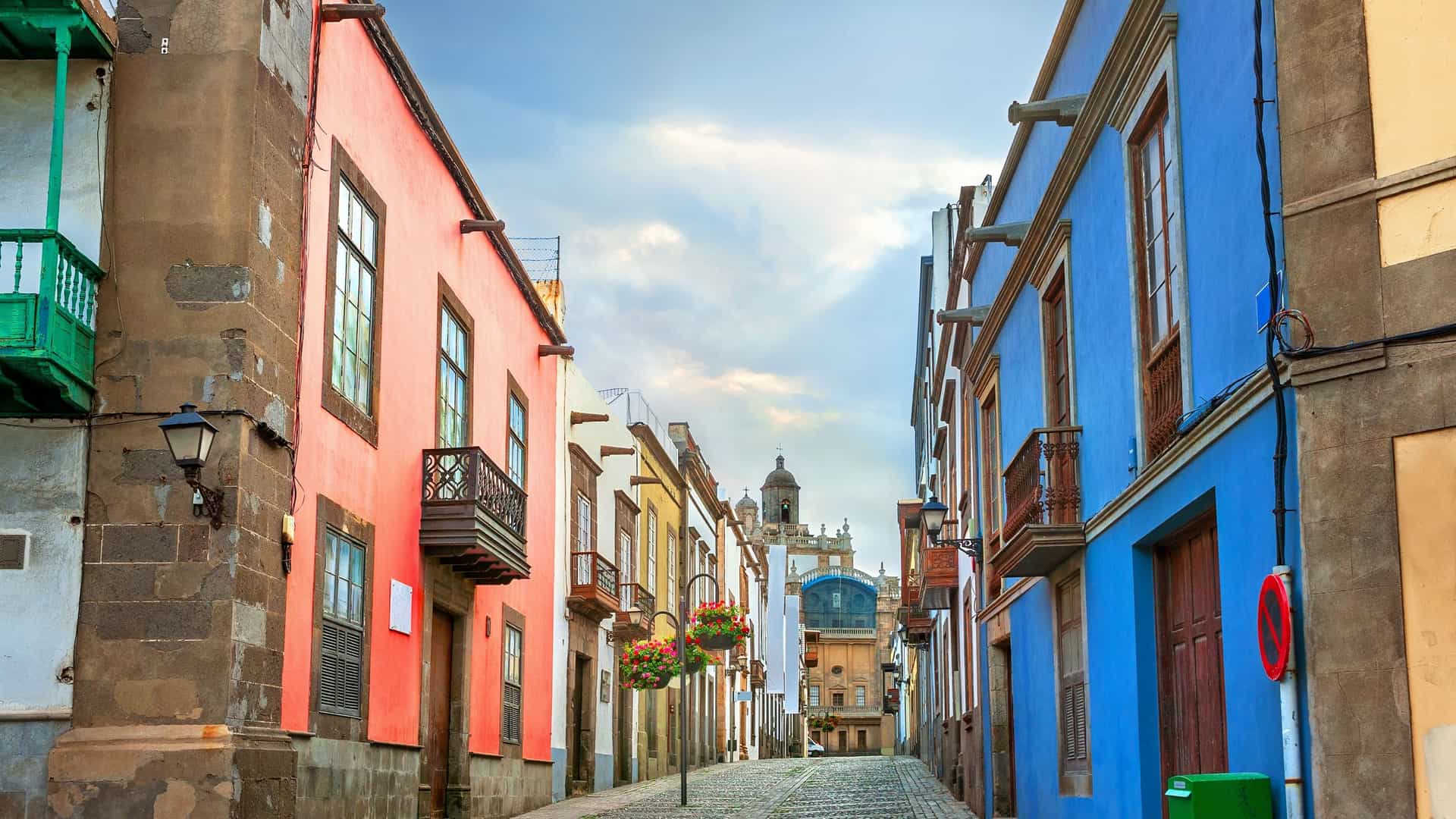 A narrow, uphill cobblestone street in the Vegueta historic district of Las Palmas, lined with brightly colored colonial-style buildings. A prominent building on the right is blue and one on the left is reddish-orange. The street leads toward a historic, light-colored building with a tower in the distance under a partially cloudy sky.