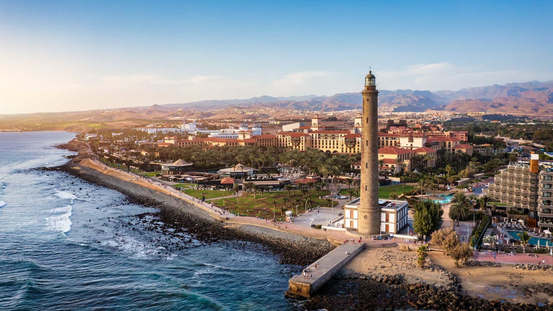 A scenic view of the Maspalomas Lighthouse, a historic 19th century landmark in Gran Canaria, Spain. The tall, cylindrical stone tower stands against a clear sky, with a smaller, traditional Canarian style building at its base. The lighthouse is located at the southern tip of the island, separating the Maspalomas beach and the famous Maspalomas Dunes from the Meloneras promenade.