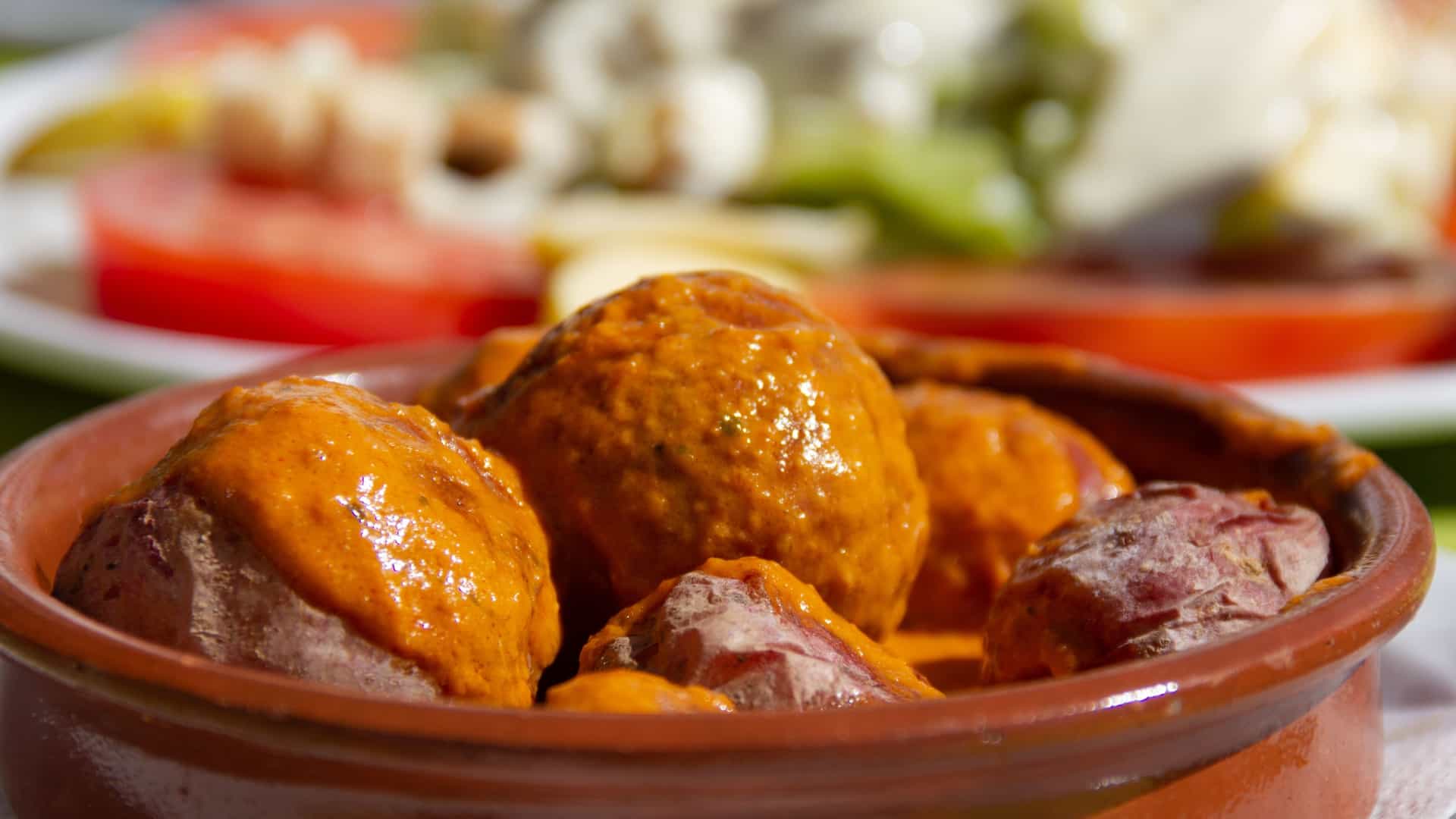 A close up, top down shot of a traditional Canarian dish called Papas Arrugadas. The small, thin skinned potatoes have a wrinkled texture and a white, salty crust from being boiled in salt water. They are served in a rustic, light colored bowl alongside two small bowls of sauce: a vibrant red, spicy mojo picón sauce and another, likely a green mojo sauce. The presentation suggests a simple, flavorful, and authentic meal.