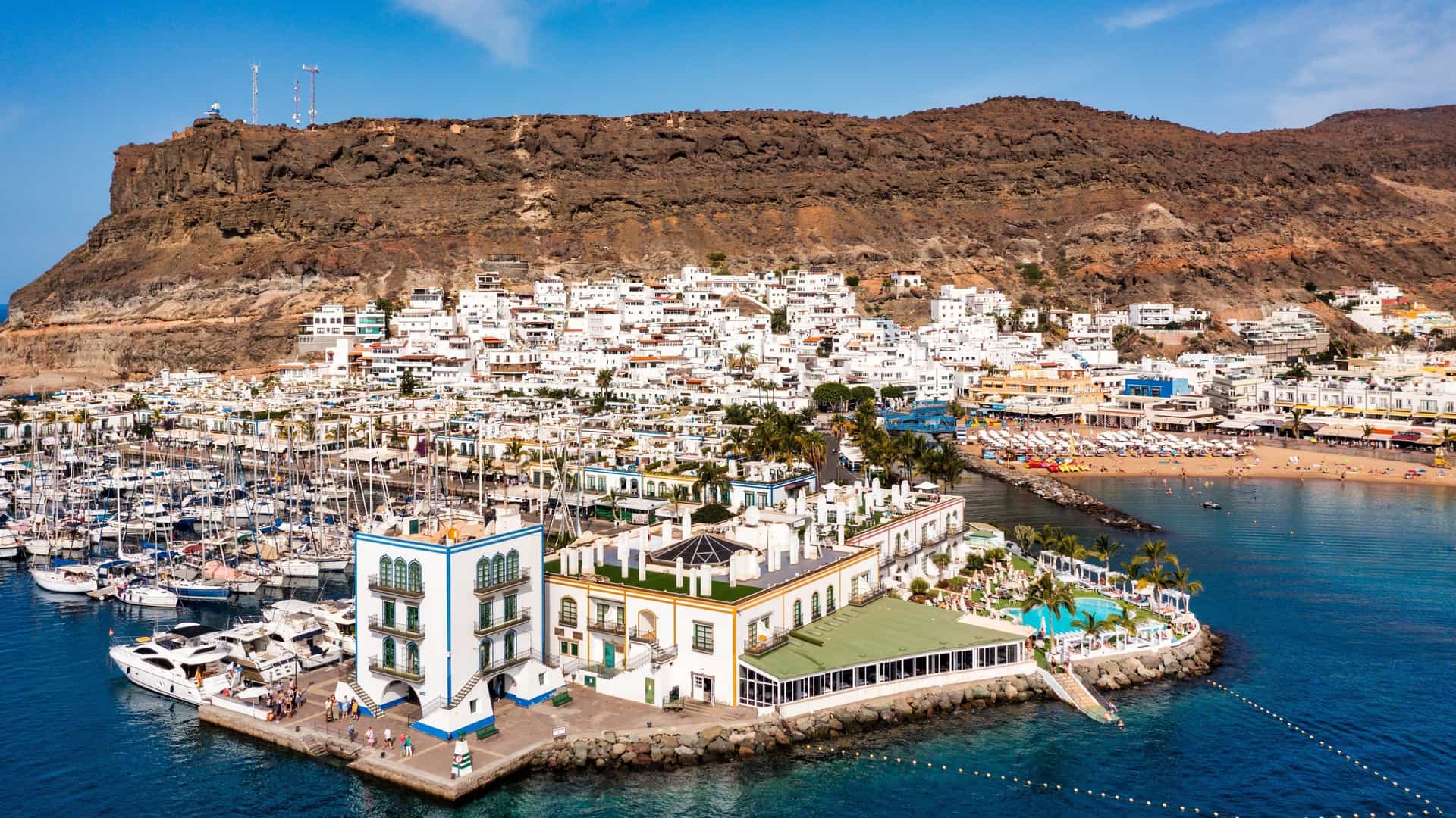 A scenic view of the man made beach at Puerto de Mogán, Gran Canaria. The calm, turquoise water of the bay is lined with golden sand, dotted with sunbathers and umbrellas. The beach is enclosed by a stone breakwater and is surrounded by the town's charming, low rise buildings with white walls and colorful trim, typical of the "Little Venice" nickname. In the background, boats are docked in the marina, and the rugged, volcanic cliffs of the island rise into the sky.