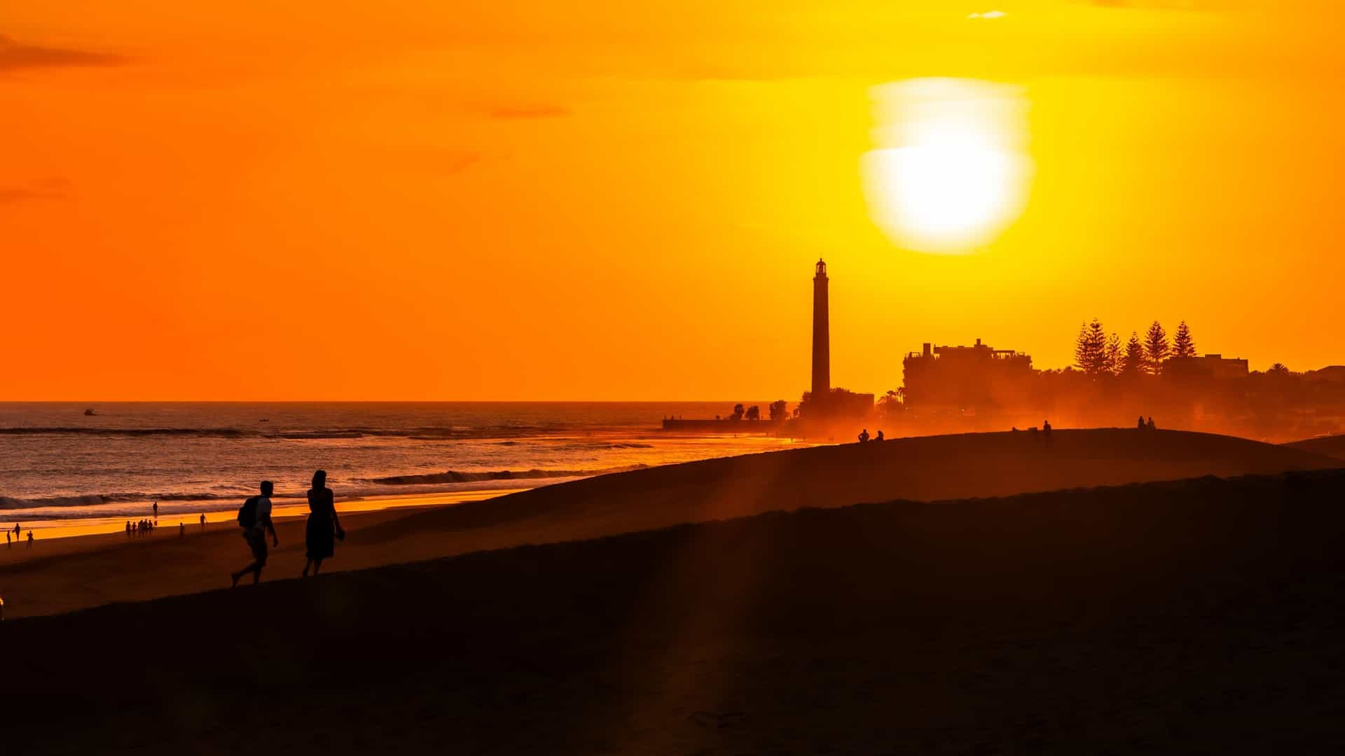 A group of tourists walking on the vast, wind sculpted sand dunes of Maspalomas in Gran Canaria. The sun is setting, bathing the dunes and the people in a warm, golden orange light. The Maspalomas lighthouse is visible in the distance, and the long shadows of the people stretch across the rippling sand. The image captures the serene beauty of the desert like landscape at dusk.