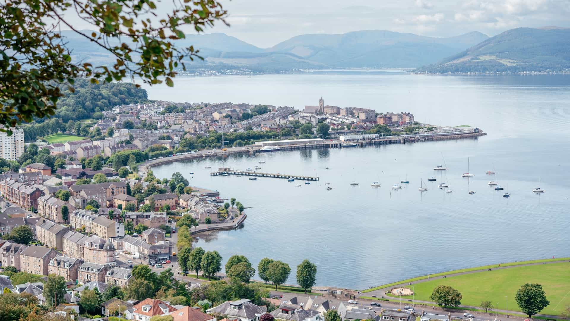 A scenic view of Gourock Bay on the Firth of Clyde in Scotland, taken on a cloudy day. The town of Gourock, with its buildings and waterfront, is visible along the shoreline. The calm waters of the bay are in the foreground, and the distant hills of the Argyll peninsula are visible across the water under a dramatic sky.