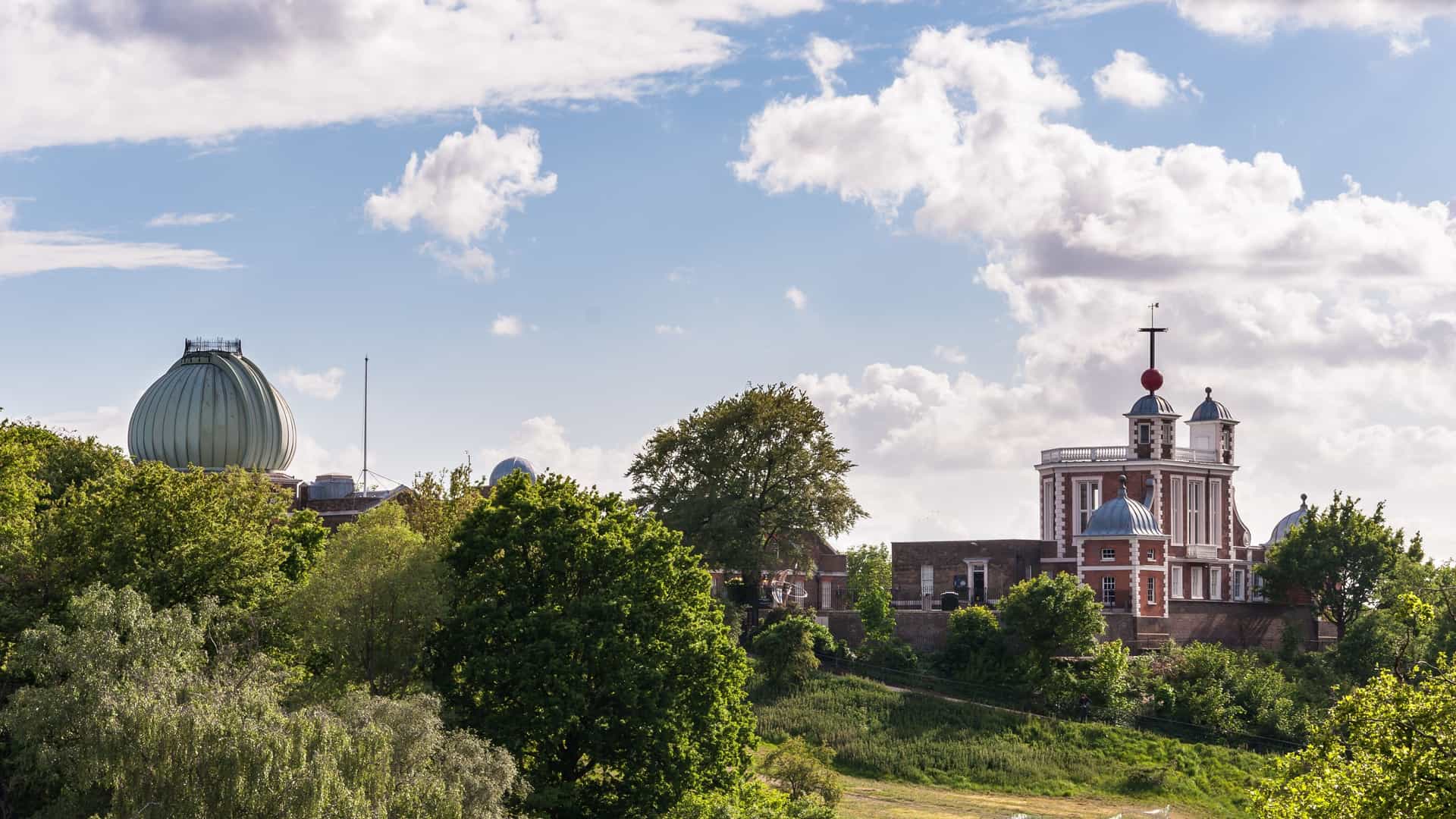 A prominent view of the Royal Observatory, a historic brick building with a distinctive dome, situated on a hill within Greenwich Park in London. The image captures the architectural details of the observatory under a bright sky, with visitors visible near the main entrance or the Prime Meridian line that runs through the courtyard.