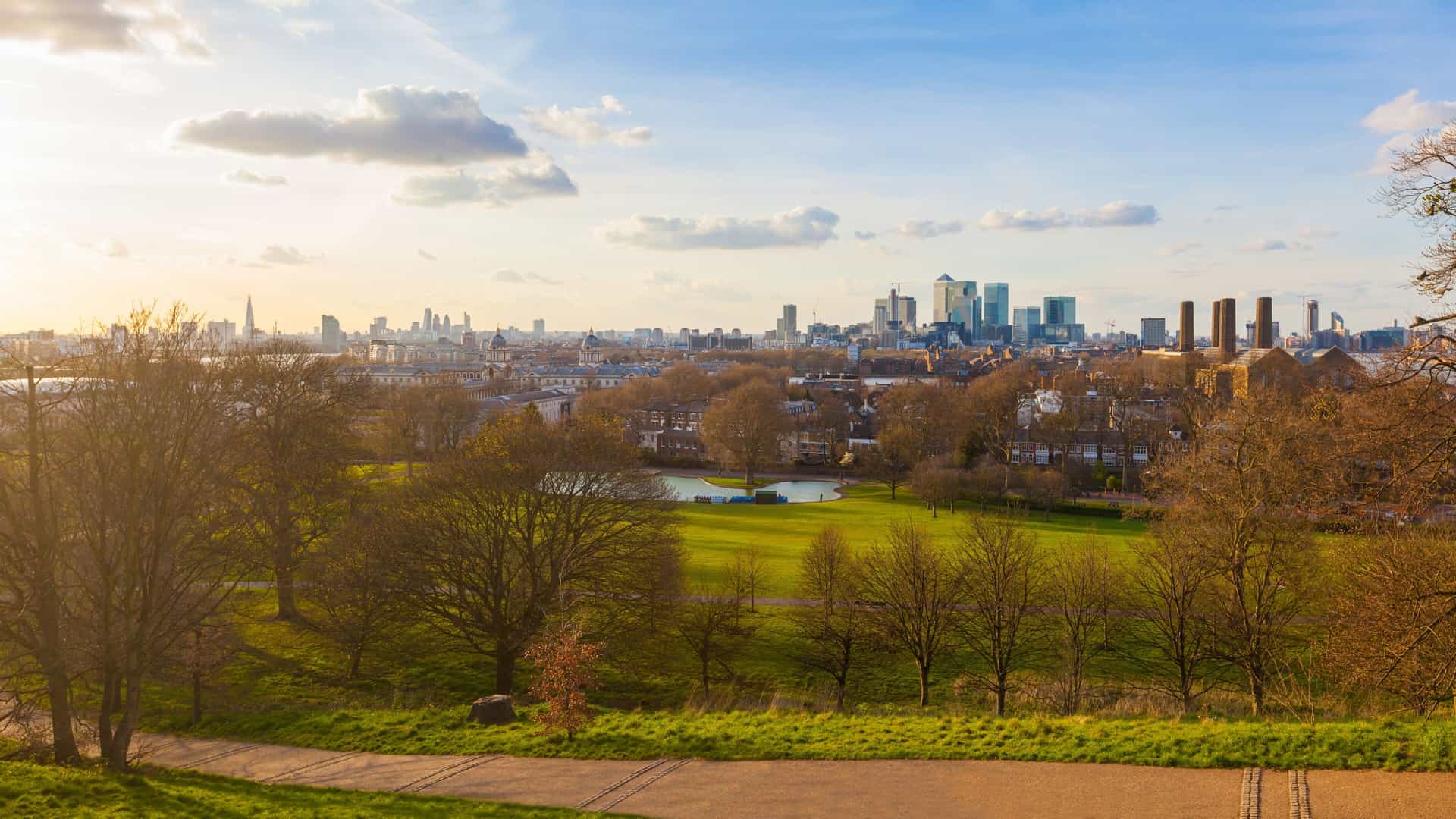 A panoramic view of the London skyline at sunset from Greenwich Park. In the foreground, the lush green park slopes down towards the Old Royal Naval College and the River Thames. The skyscrapers of Canary Wharf dominate the mid ground, while the distant buildings of the City of London are visible on the horizon, all silhouetted against a sky with golden and pink hues.