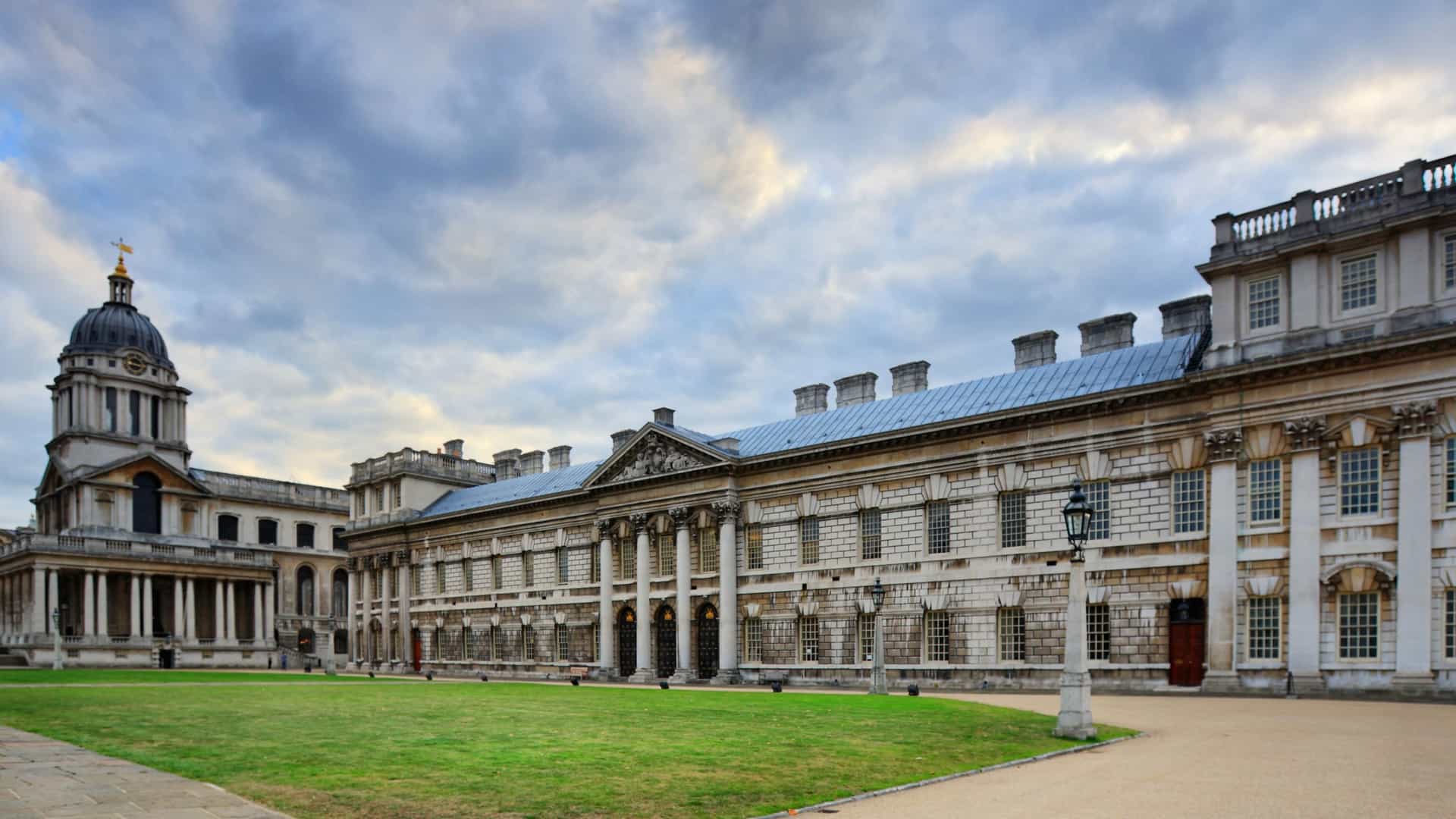 The stately, classical facade of the National Maritime Museum in Greenwich, London, is captured on a bright day. The large, historic white stone building stands prominently with a wide, manicured lawn in the foreground. The image emphasizes the museum's grandeur as the leading maritime museum of the United Kingdom, set against a clear sky.