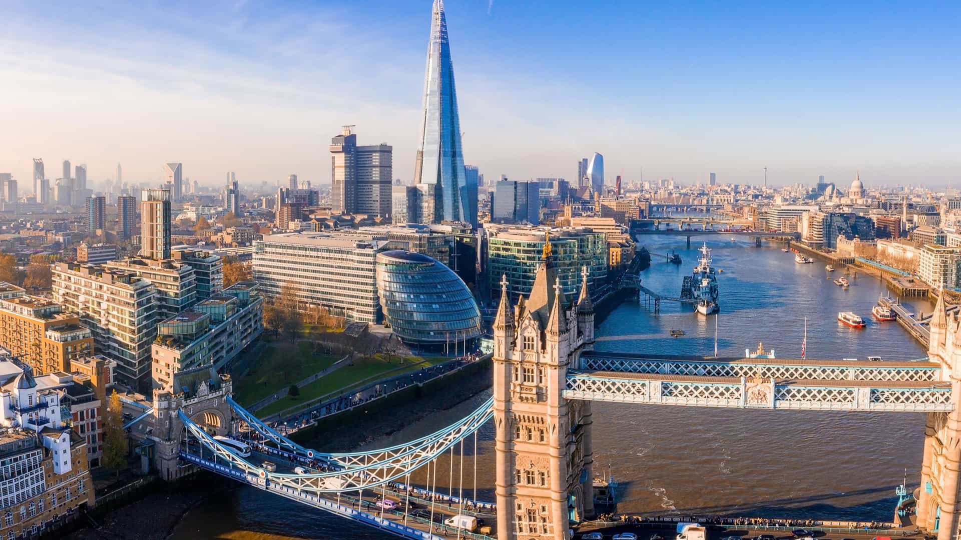 A stunning panoramic view of the Thames River and the London skyline at dusk. The water reflects the city lights, and landmarks such as the Tower Bridge, The Shard, and other skyscrapers are visible against the colorful evening sky.