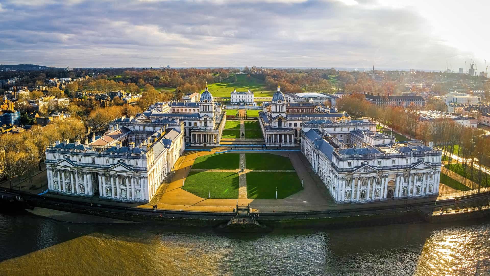 An aerial view of the Old Royal Naval College in Greenwich, London, showing its baroque architecture and two prominent domes. The River Thames curves around the college's waterfront, with the skyscrapers of the Canary Wharf business district visible across the river in the background. The college is surrounded by green lawns and is situated next to other notable landmarks, including the Queen's House.
