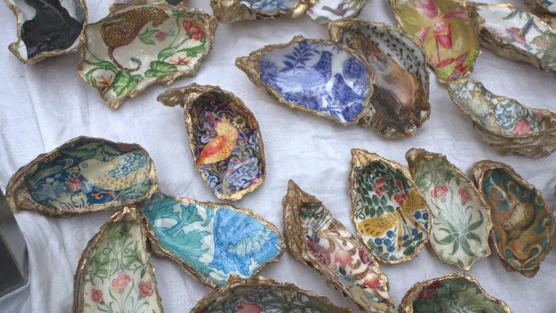 A close-up view of an array of decorative, colorful painted seashells and oyster shells displayed for sale at a stall in Greenwich Market, London. The shells are painted with detailed miniature scenes, floral patterns, and abstract designs in vibrant colors, arranged closely together on a surface to showcase the handcrafted artistry.