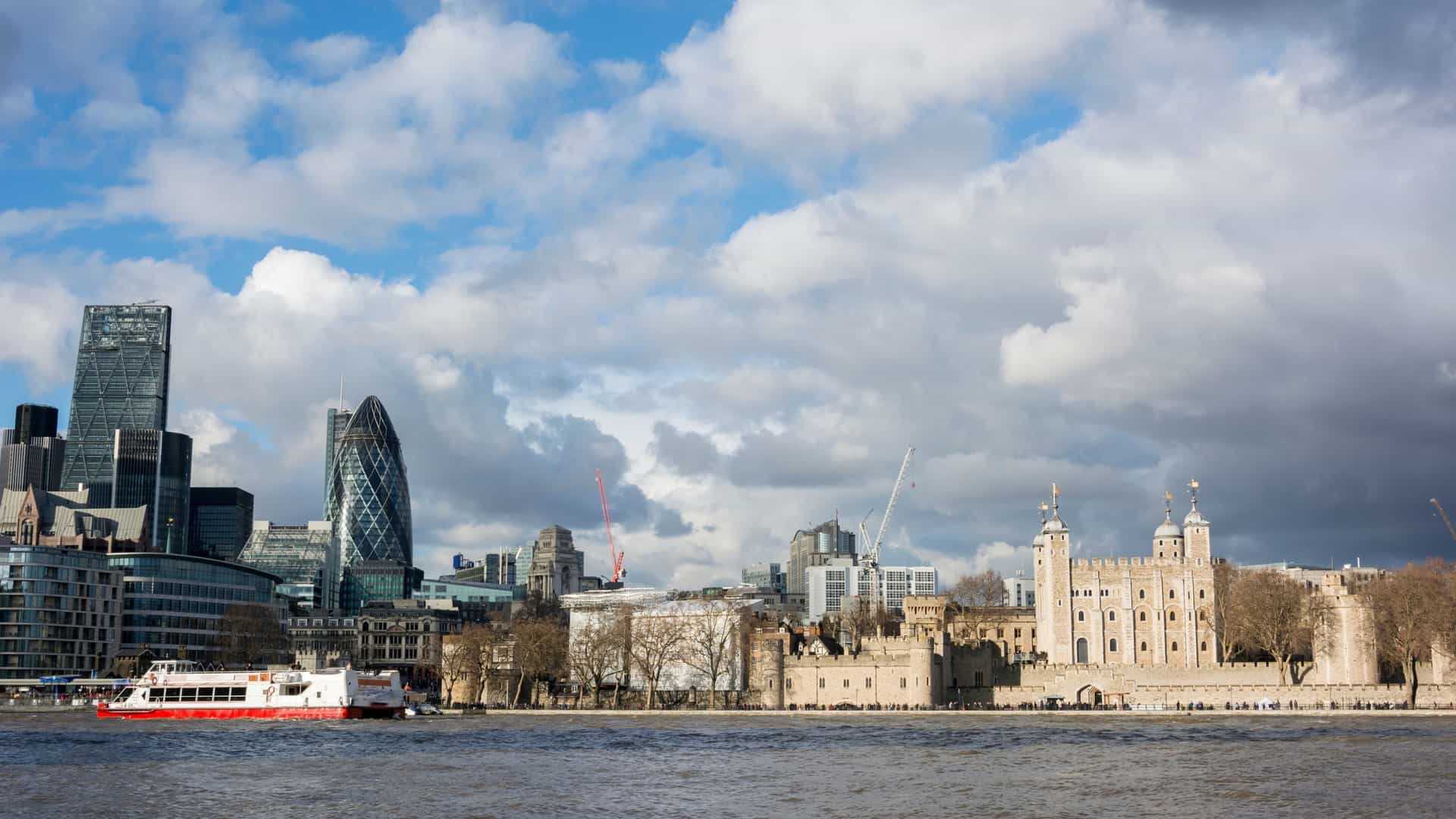 A sweeping, panoramic view of the Central London skyline, dominated by the River Thames in the foreground, features a prominent tour boat or ferry, suggesting a River Thames Cruise to Central London. Dramatic, dark storm clouds hang low and heavy over the cityscape, casting deep shadows that contrast with the illuminated, recognizable buildings of the city's financial district and iconic landmarks.