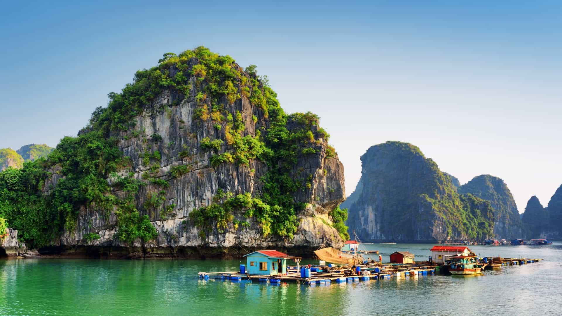 A serene view of a floating fishing village in Ha Long Bay, Vietnam. Numerous small, brightly colored floating houses and fishing boats are clustered together on the calm, emerald green water. The village is surrounded by the iconic, towering limestone karsts of the bay, with a dramatic mountainous landscape in the background under a hazy sky.