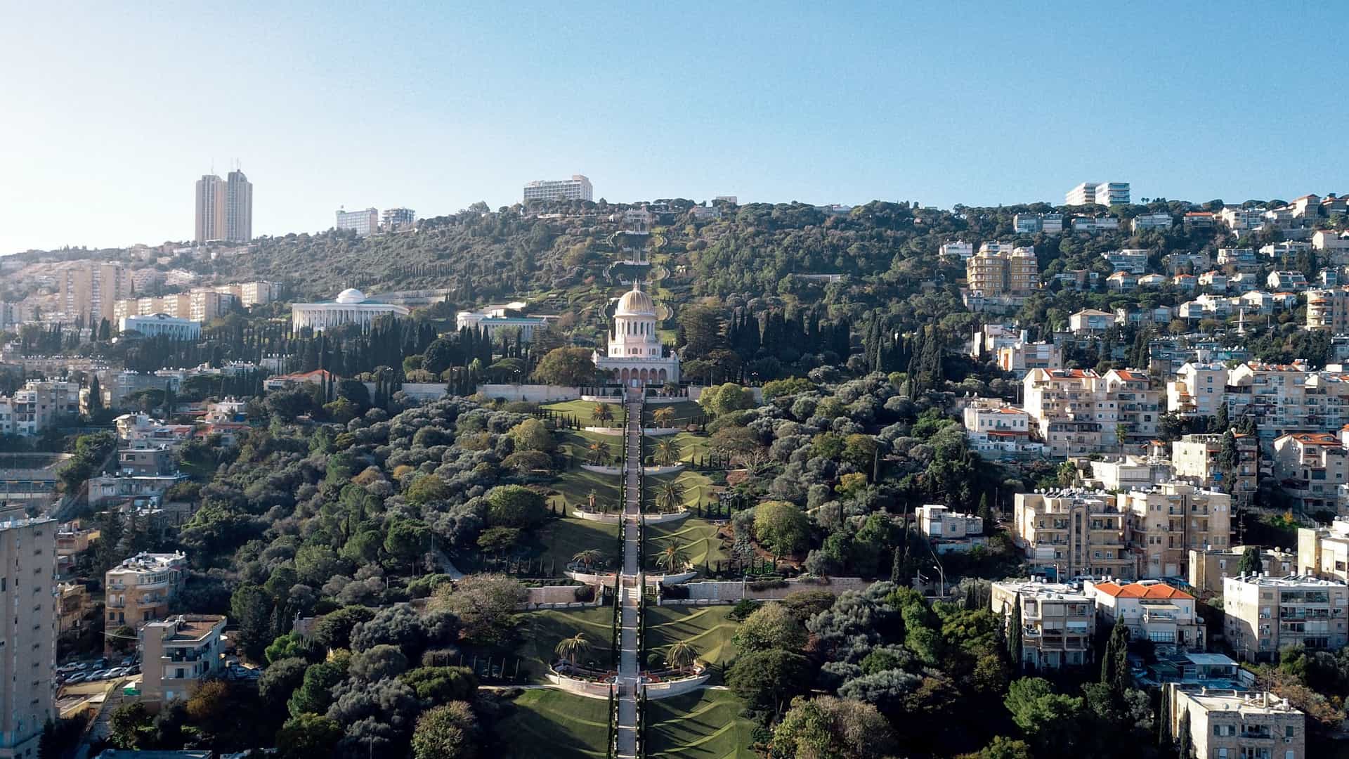 An aerial view of the Baháʼí Gardens in Haifa, Israel, showing the nineteen meticulously manicured, concentric terraces with perfectly symmetrical geometric patterns, leading down from the golden domed Shrine of the Báb, with a view of the city of Haifa and the Mediterranean Sea in the background.