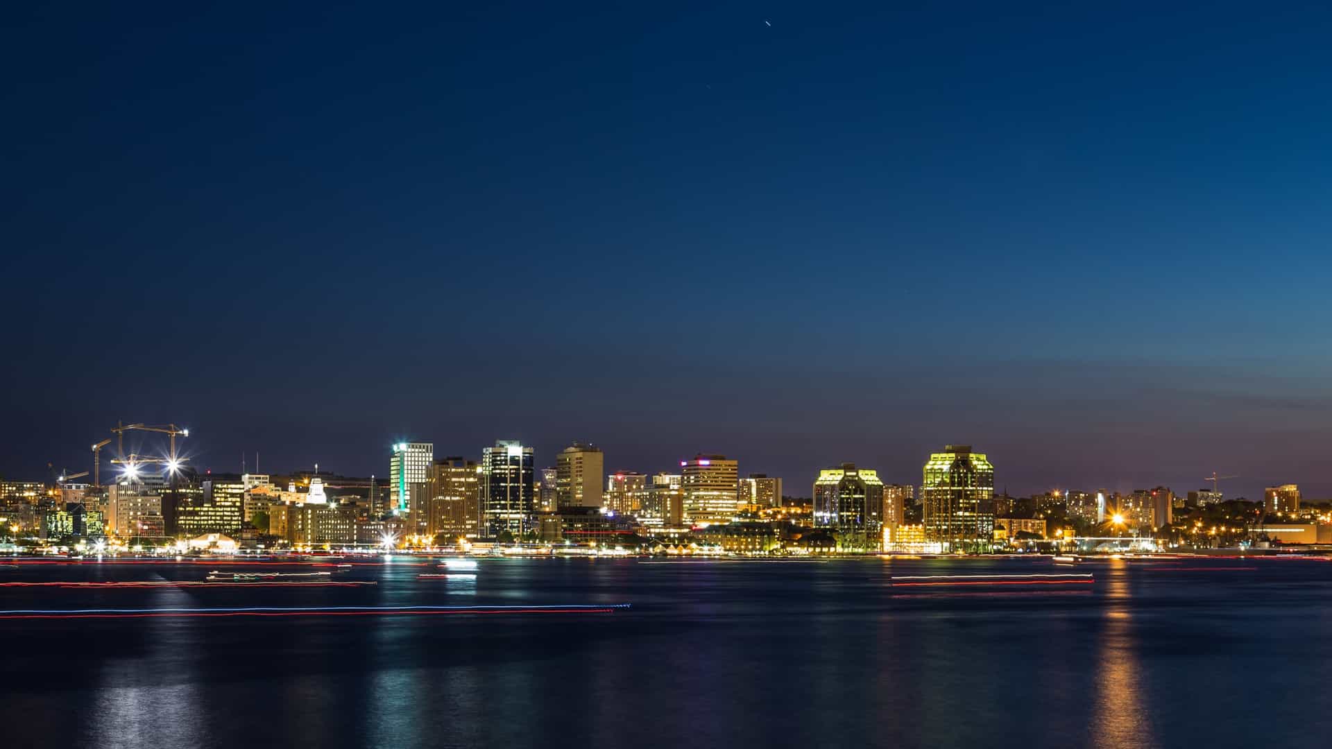 A long exposure photograph of the Halifax waterfront at night. The city's skyline is illuminated, with lights reflecting on the calm harbor water. There are streaks of light, a "trail of boats," created by the movement of boats on the water, and the waterfront is lined with illuminated buildings.