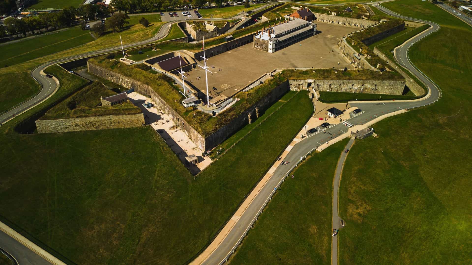 An aerial view of the Halifax Citadel National Historic Site in Nova Scotia. The star shaped, stone fortification is visible atop Citadel Hill, surrounded by green grass. The image also shows the surrounding downtown Halifax area and the harbor in the distance.