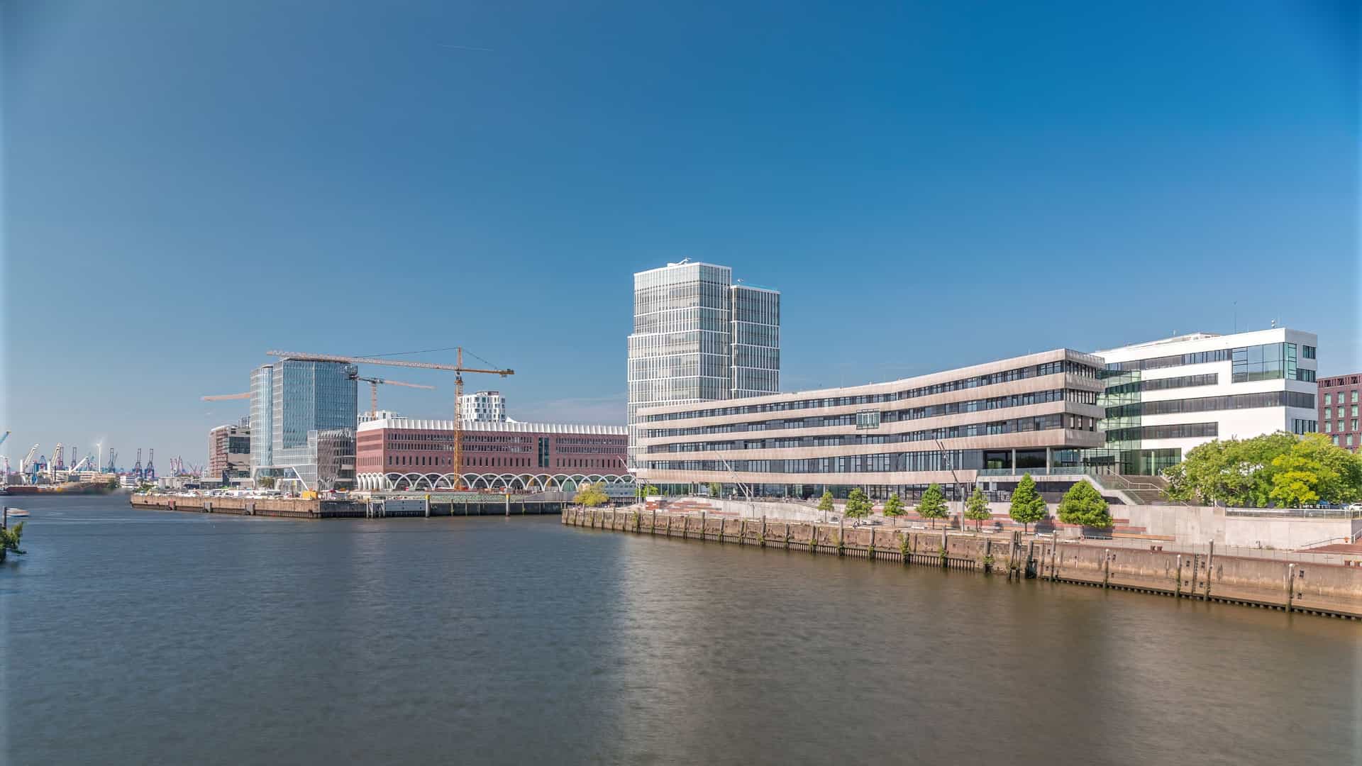 A nighttime timelapse of HafenCity University in Hamburg, Germany. The modern architectural building is prominently featured, illuminated against the dark sky. The shot includes the waterfront skyline, construction cranes, and the blurred trails of light from passing vehicles and boats, characteristic of a long exposure photograph.