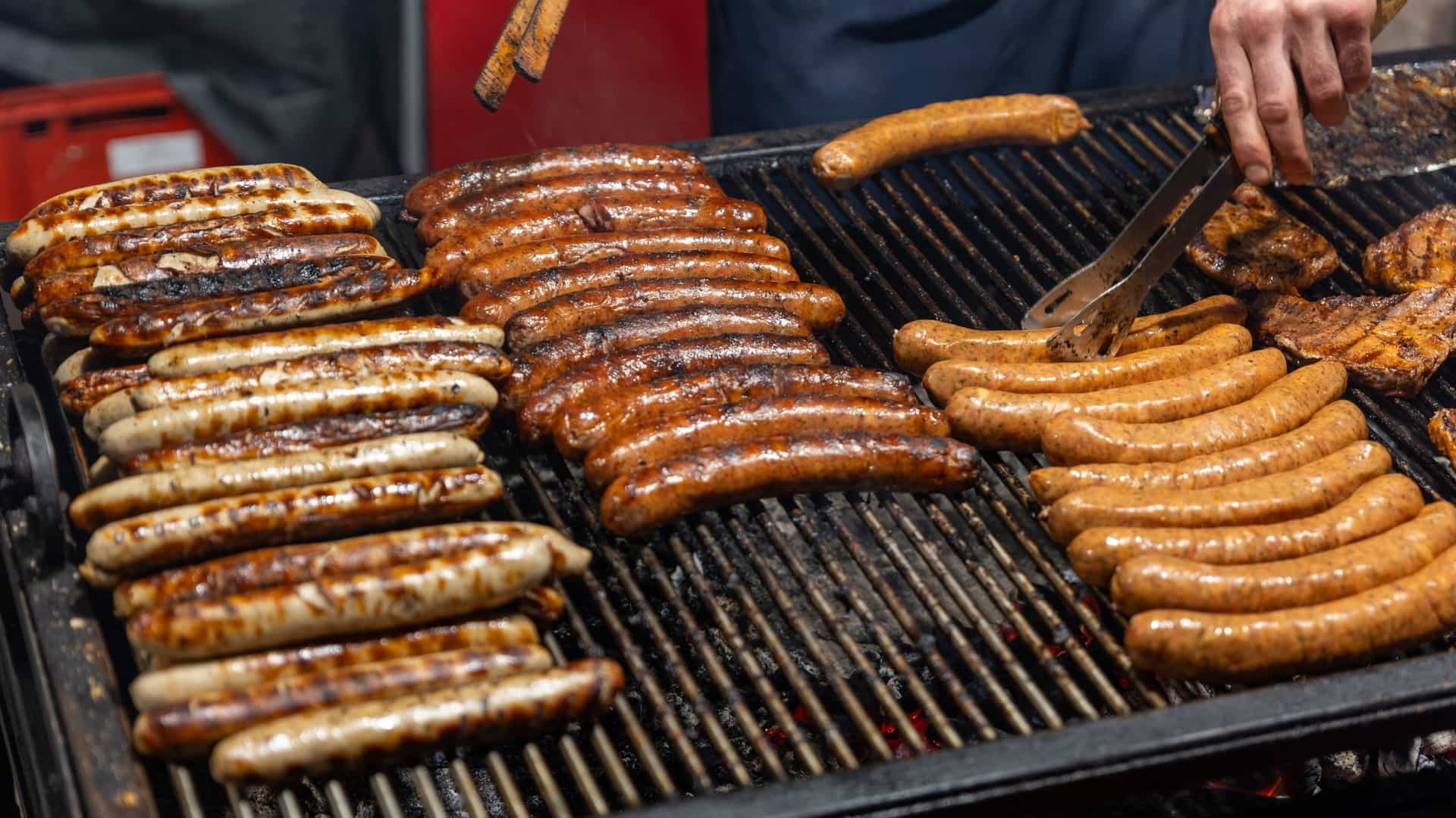 A close up view of several juicy bratwurst sausages and meat steaks sizzling on a hot grill at a German street food market. The food is shown up close, highlighting the texture and moisture of the meats as they cook.