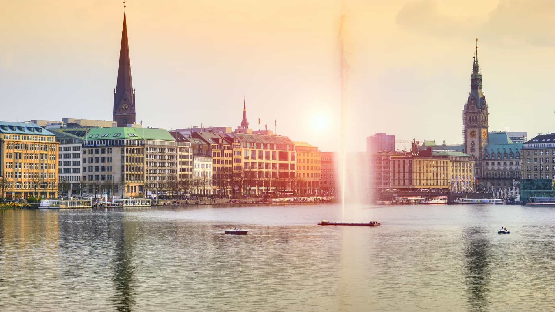 A scenic photograph of the Alster Lake in Hamburg, Germany, at sunset. The sky is filled with warm, golden light, casting a vibrant reflection on the calm water. The silhouette of Hamburg's skyline, with its prominent buildings and spires, is visible in the background, creating a picturesque view.