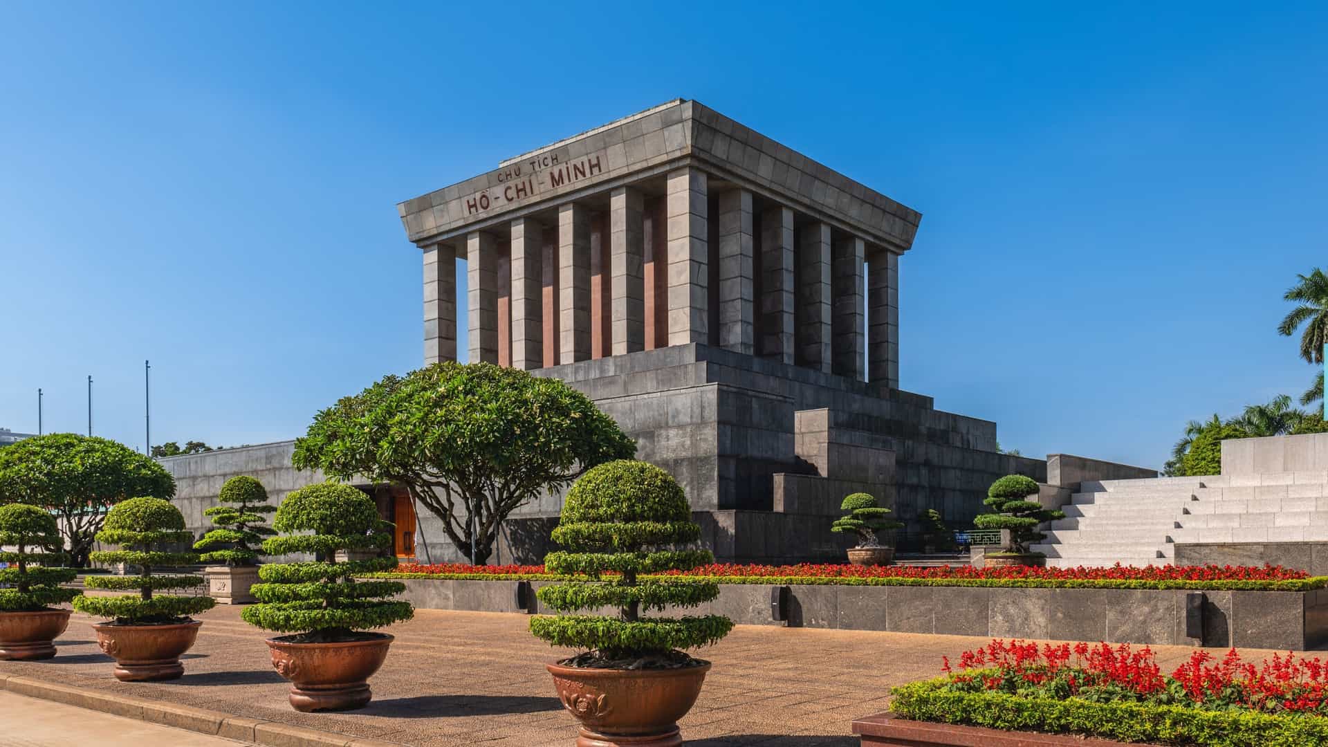 A photograph of the Ho Chi Minh Mausoleum in Hanoi, Vietnam. The monumental, cube like building is made of dark gray granite and features a three tiered base and a stepped roof, with large, square pillars supporting the front. The inscription "CHỦ TỊCH HỒ CHÍ MINH" is visible on the upper part of the facade. The mausoleum stands in a large, paved square with a clear sky above.