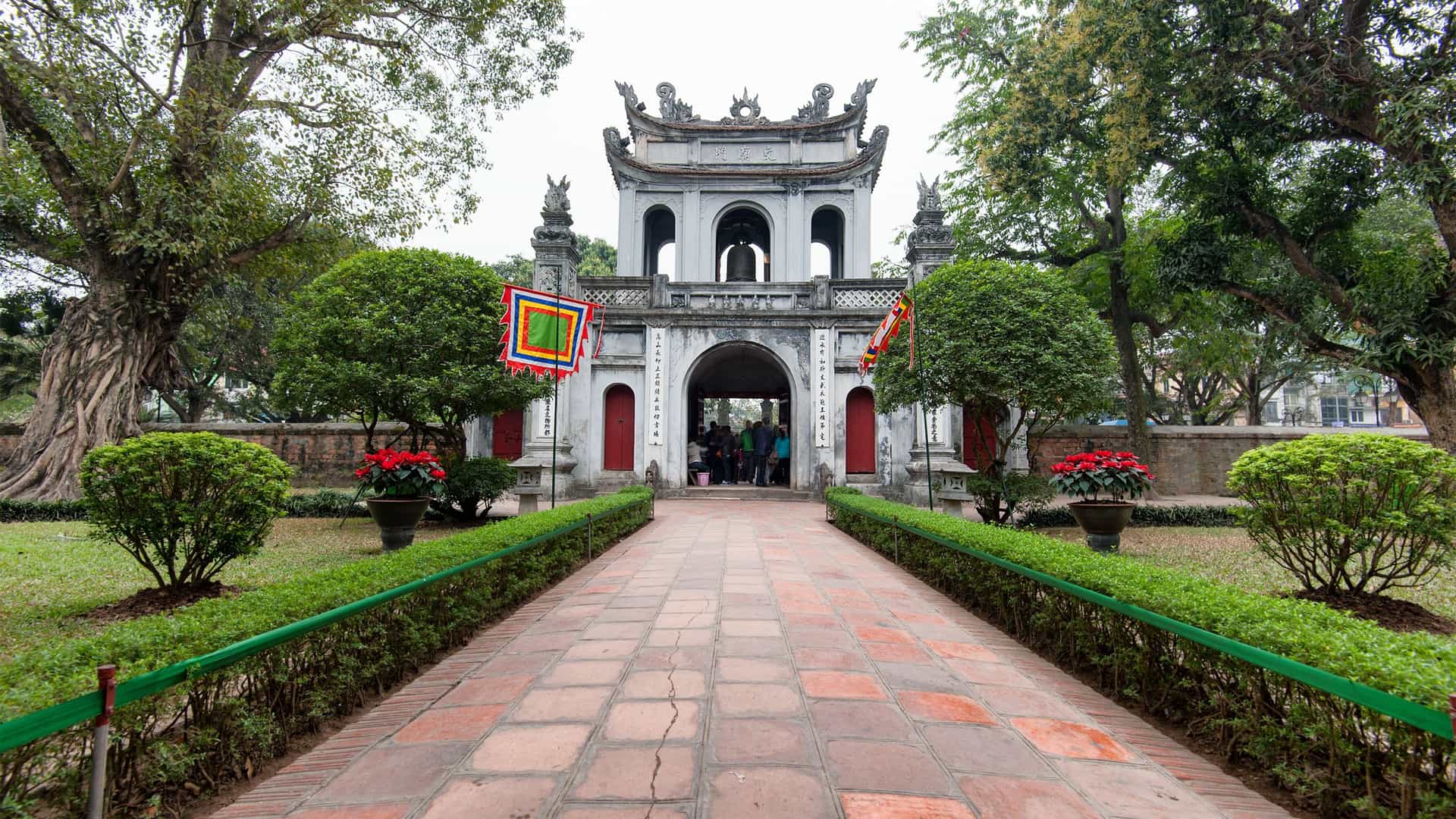 A photograph of the main entrance gate of the Temple of Literature in Hanoi, Vietnam. The gate, known as the Great Portico or Van Mieu Gate, is a two story structure with three arched doorways and a red tiled roof. The largest central doorway is flanked by two smaller side doors. Four tall, decorative pillars stand in front of the gate, and lush green trees are visible within the temple complex.
