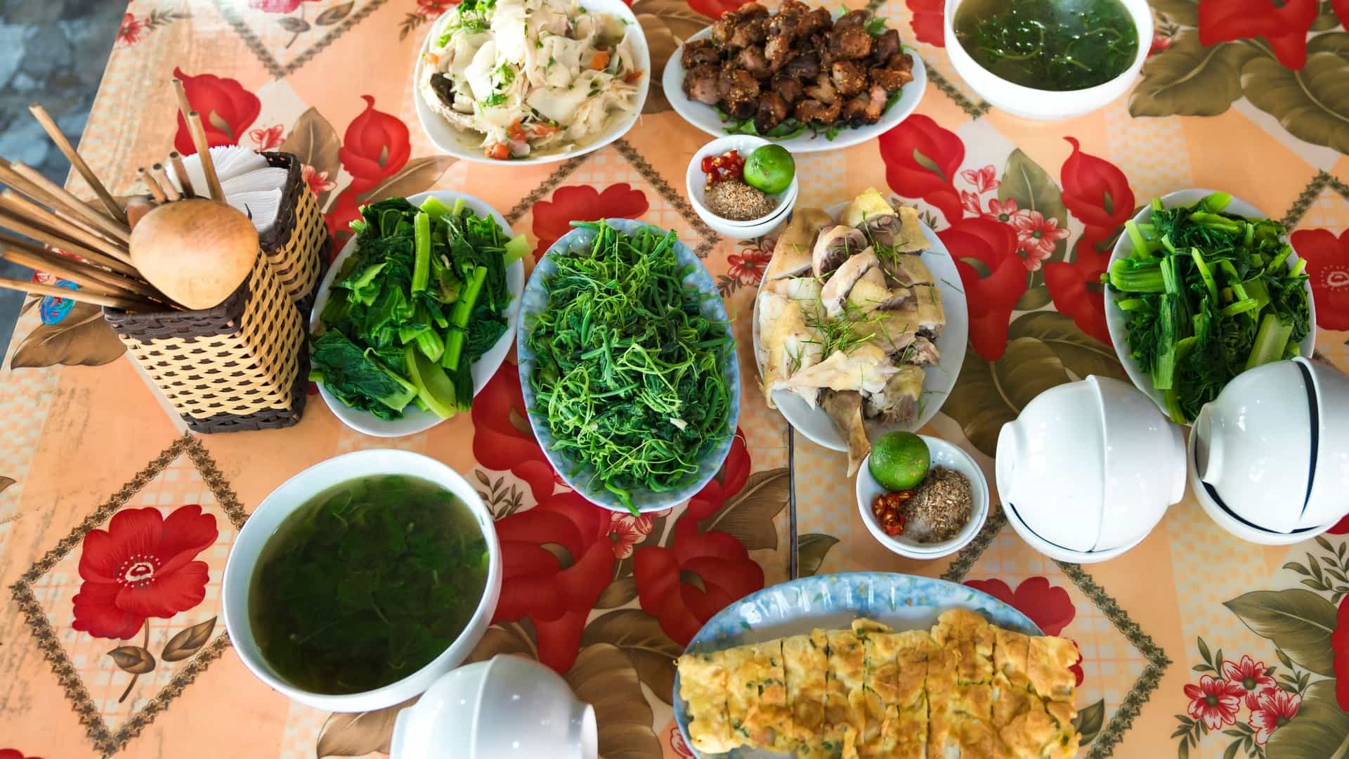 A high angle shot of a Vietnamese family style lunch set, served on a round bamboo tray. The meal includes a variety of dishes in separate bowls, such as steamed white rice, a salad, sliced boiled chicken, fried pork, and boiled vegetables. A plate of fried eggs is also visible.