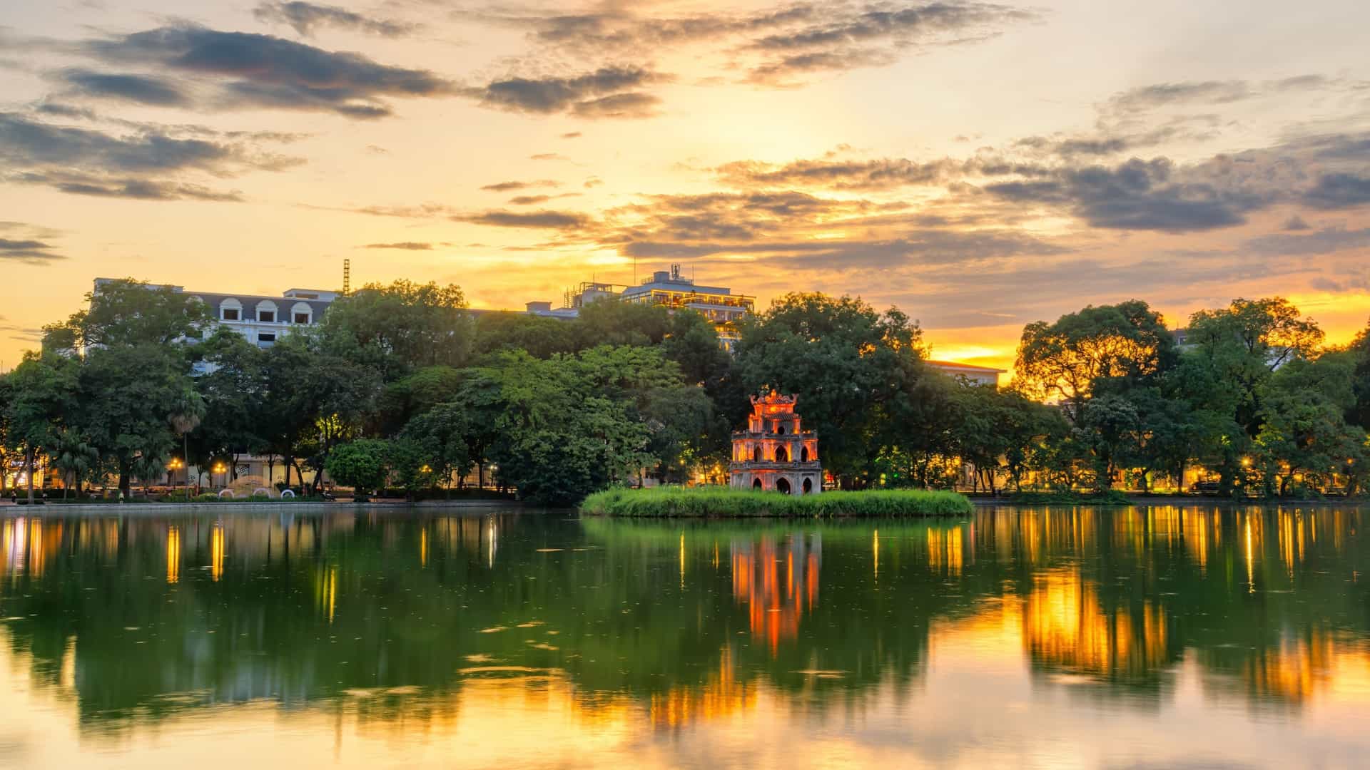 A scenic photograph of Hoan Kiem Lake in Hanoi, Vietnam, also known as Sword Lake. The image captures the iconic Turtle Tower (Thap Rua) on its small island, centered in the tranquil green water. The lake is surrounded by lush trees, with modern buildings and a clear sky in the background, showing the contrast between nature and the urban landscape.