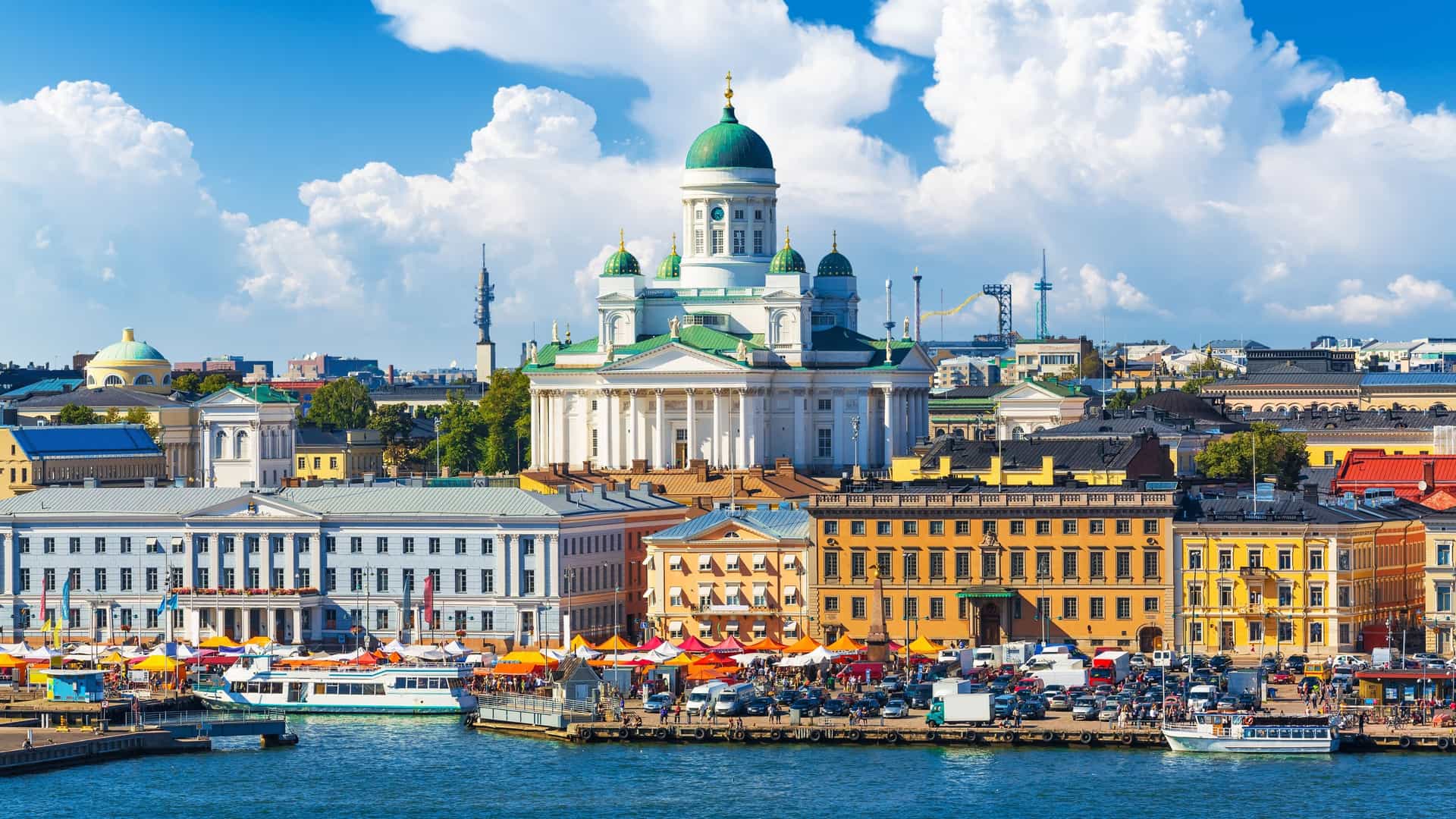 A scenic panoramic photograph of the Market Square (Kauppatori) in Helsinki, Finland, on a summer day. The image shows the bustling open air market with vendors' tents and people walking around. In the foreground, there are boats docked at the Old Town pier, and in the background, the cityscape of Helsinki, including the Helsinki Cathedral, is visible under a clear sky.