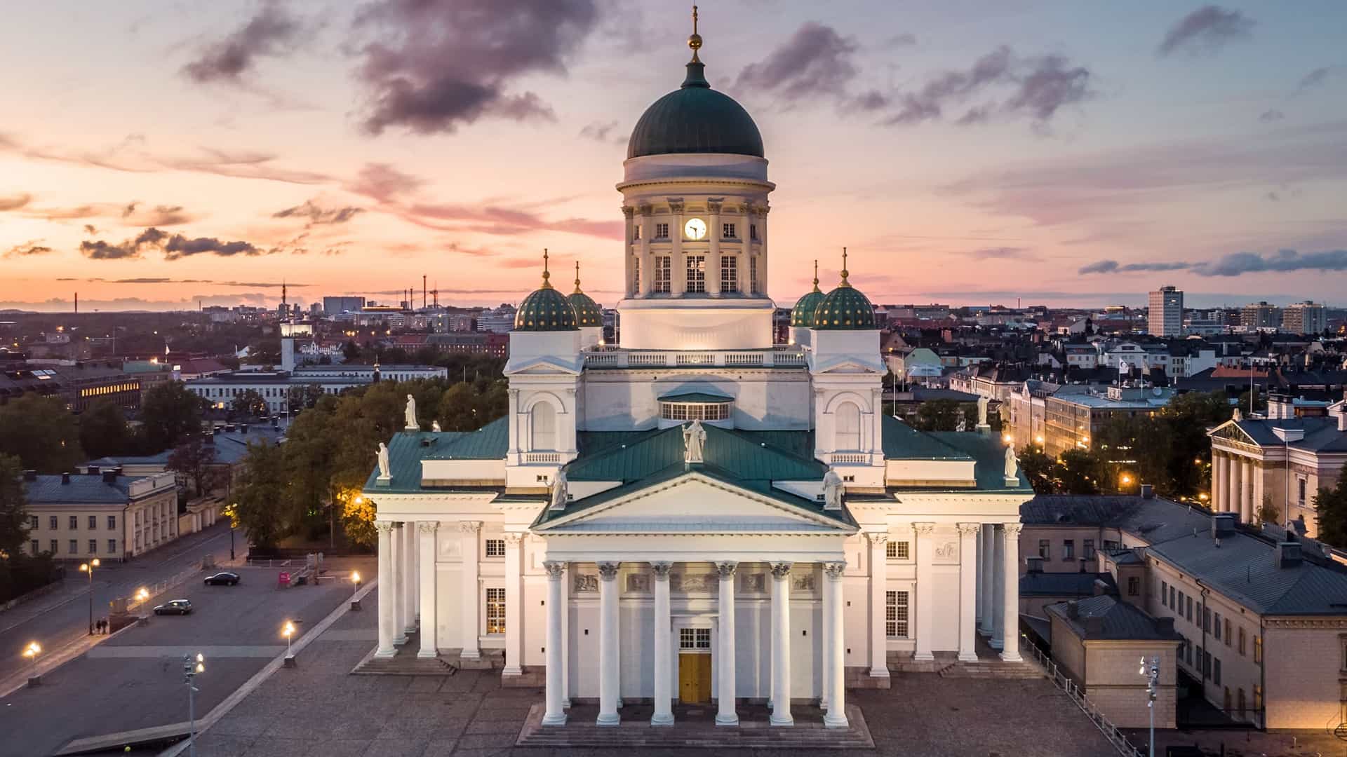 An aerial view of Helsinki, Finland, during the autumn season. The photograph captures a clear view of the city's waterfront, with buildings, streets, and trees with colorful fall foliage. The Helsinki Cathedral is prominently visible in the image, along with other landmarks, under a bright, sunny sky.