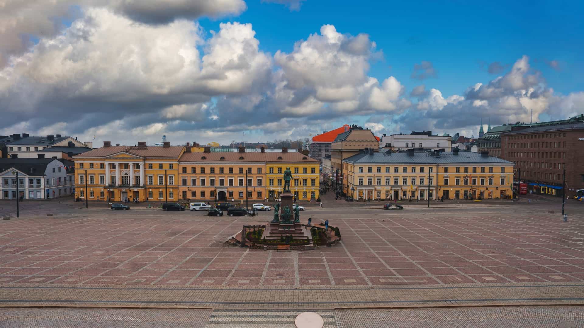 A photograph of Helsinki's Senate Square, showcasing its Neoclassical architecture. In the center of the square stands the bronze statue of Alexander II on a granite pedestal. In the background, the grand, white Helsinki Cathedral with its green domes and golden crosses dominates the scene, while other historic buildings, such as the Government Palace and the main building of the University of Helsinki, frame the square.
