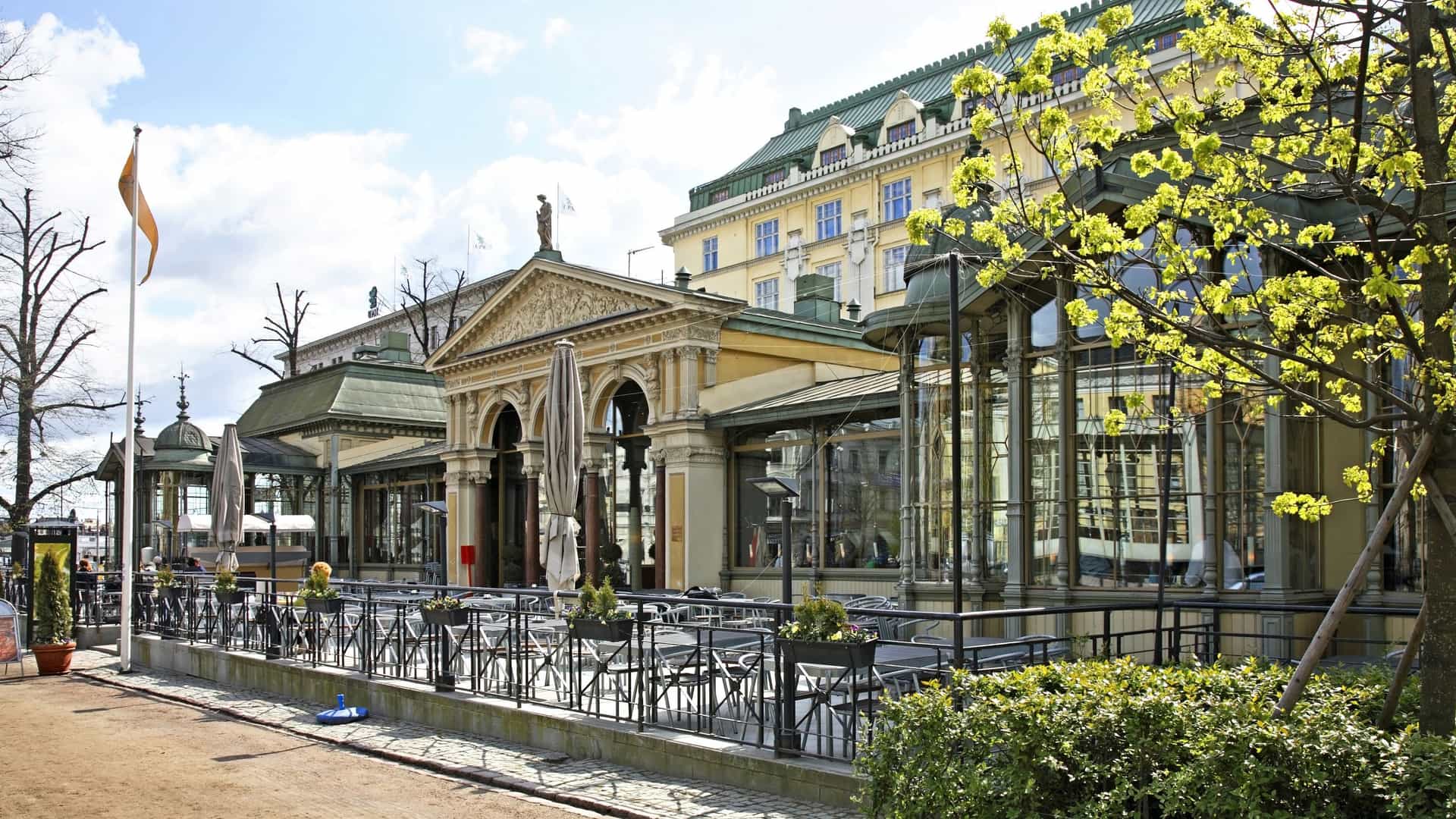 A photograph of Esplanadi Park in Helsinki, Finland. The image shows a wide, tree lined avenue with lush green lawns and colorful flowerbeds. People are seen strolling along the pathways and relaxing on benches, enjoying the sunny day. Historic buildings, including the Swedish Theatre and Restaurant Kappeli, are visible in the background, framing the park.