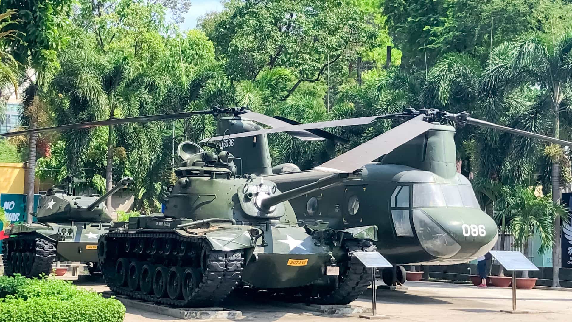 An American-made Boeing CH-47 Chinook cargo helicopter on outdoor display at the War Remnants Museum in Ho Chi Minh City (Saigon), Vietnam. The massive helicopter is captured from a high angle, showcasing its green fuselage, twin rotors, and the context of the museum's forecourt, where other military vehicles are exhibited.
