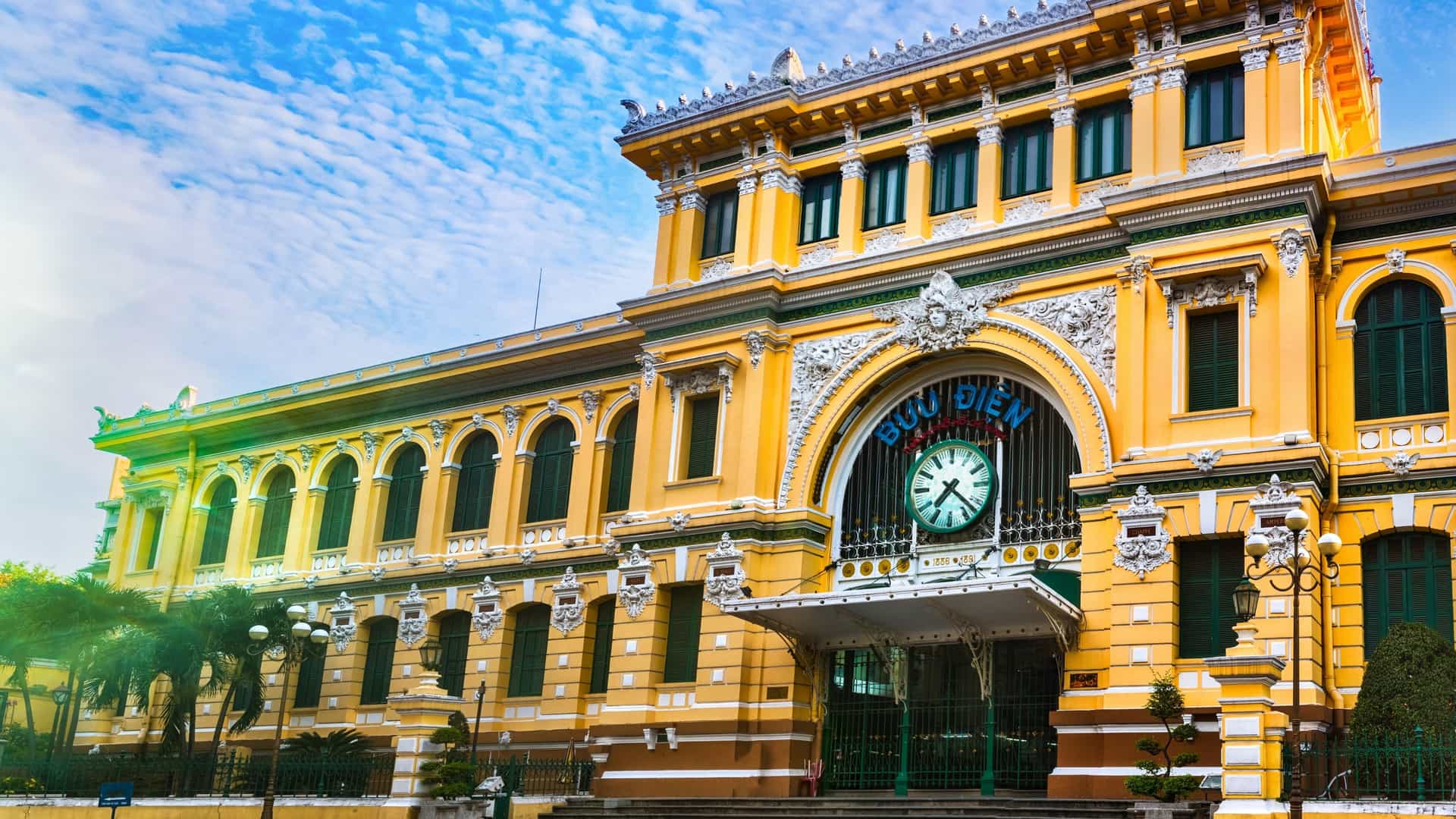 A photograph of the Saigon Central Post Office in Ho Chi Minh City, Vietnam. The French colonial style building is prominently featured with its distinct yellow facade, arched windows, and a large clock above the main entrance. The image shows the exterior of the historic building, which is both a tourist attraction and a functioning post office.