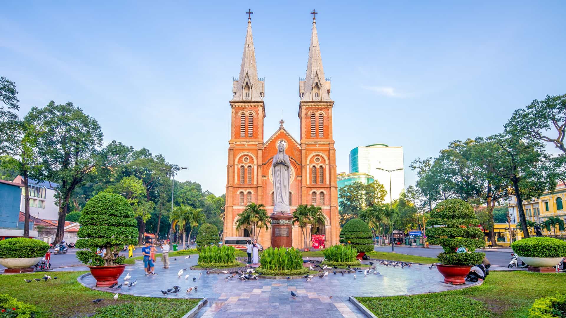 A photograph of the Notre Dame Cathedral Basilica of Saigon in Ho Chi Minh City, Vietnam. The image shows the front of the French colonial era church, with its distinctive red brick facade, large archways, and two towering bell spires. A statue of the Virgin Mary is visible in the foreground, and the sky is a bright blue. The cathedral stands as a historical and architectural landmark in the bustling downtown area.