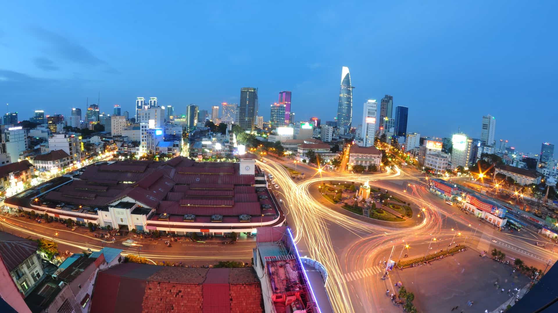 A photograph of the Ben Thanh Market in Ho Chi Minh City, Vietnam. The image shows the main entrance of the historic market, which is characterized by a prominent clock tower and French Indochinese architecture. The surrounding area is bustling with traffic and people, reflecting the market's role as a major hub in the city.