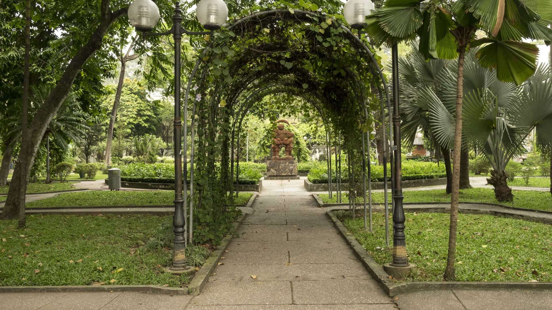 A wide-angle view of the central square and gardens of Tao Dan Park in Ho Chi Minh City, Vietnam. The photograph likely captures the park's tranquil atmosphere, featuring lush green lawns, mature trees, and perhaps a view of the Cham tower replica or other monuments found within this large urban green space.