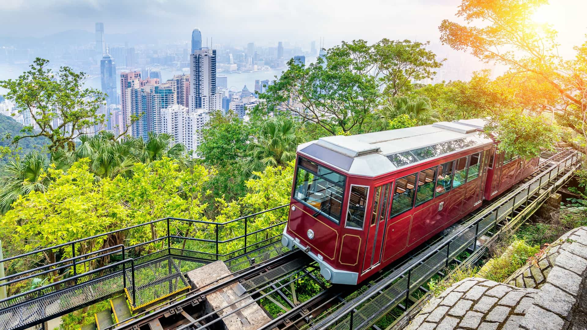 A view from inside the Victoria Peak Tram in Hong Kong, looking out at the city's skyline. The tram's red exterior and the steep angle of the railway tracks are visible. Through the large window, the dense collection of skyscrapers in Hong Kong is visible, along with the lush greenery on the mountain slope.